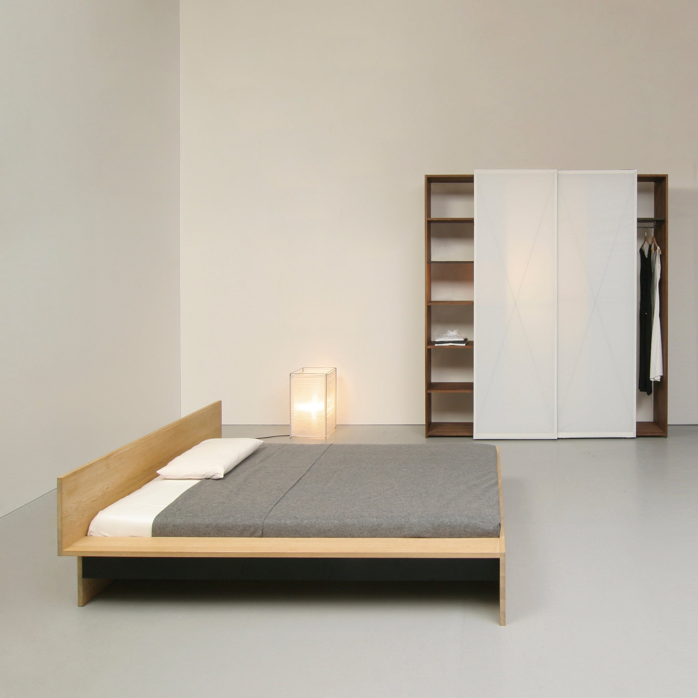 A minimalist bedroom highlights the customizable Sanktjohanser Dinadrei Bed with gray bedding. Soft light from a floor lamp complements a wardrobe featuring sliding doors and open shelving, creating a clean, modern look.