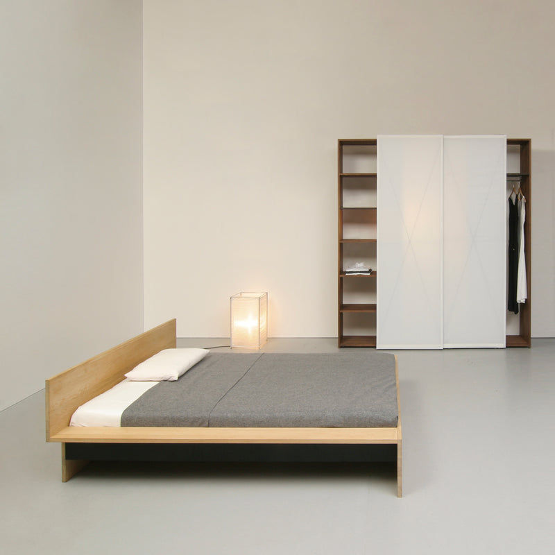 A minimalist bedroom highlights the customizable Sanktjohanser Dinadrei Bed with gray bedding. Soft light from a floor lamp complements a wardrobe featuring sliding doors and open shelving, creating a clean, modern look.