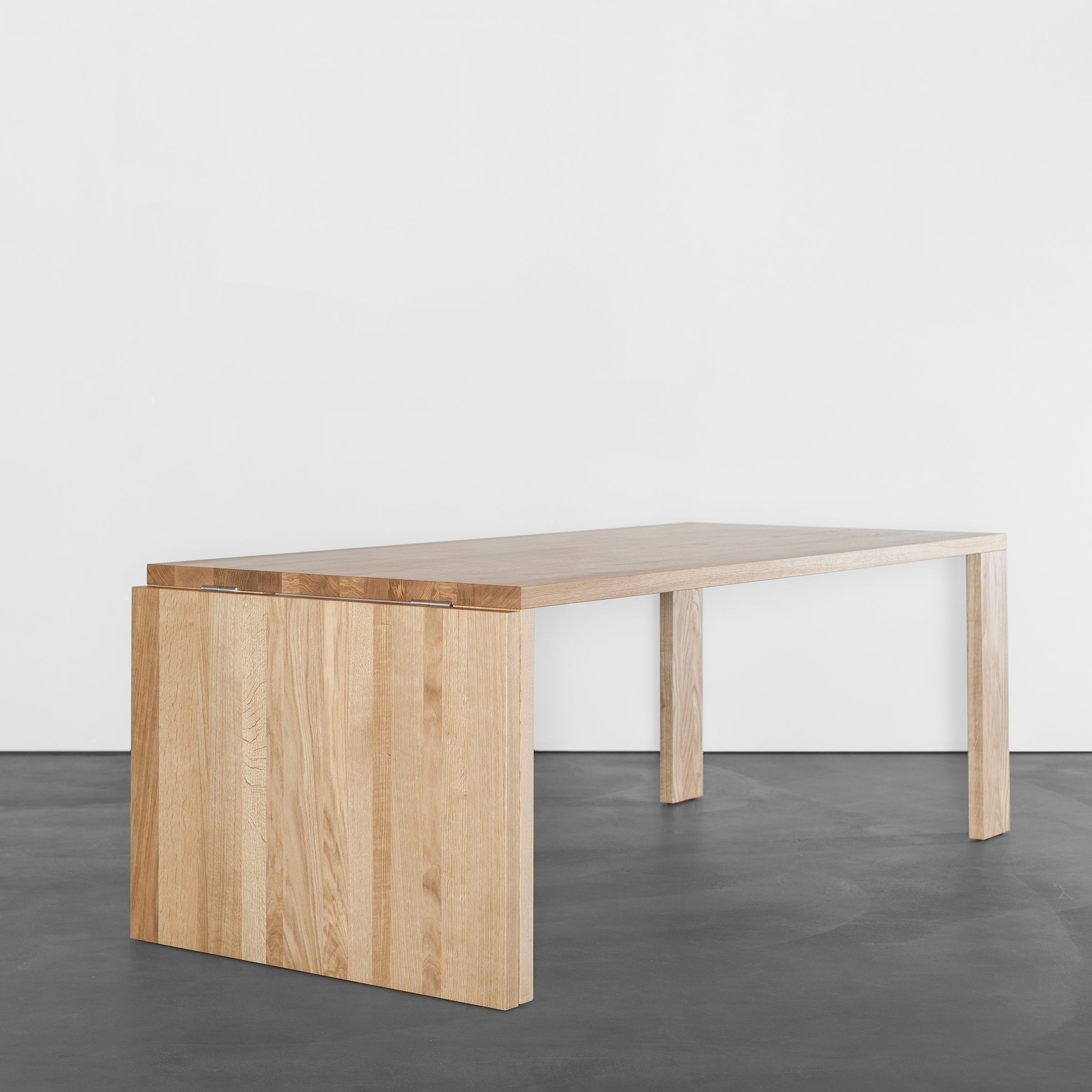 The Sanktjohanser Alphabet Folding Table, with its minimalist design and clean, angular lines, stands on a dark floor. Available in custom sizes, its light wood finish contrasts against a plain white background to highlight its simple and modern aesthetic.