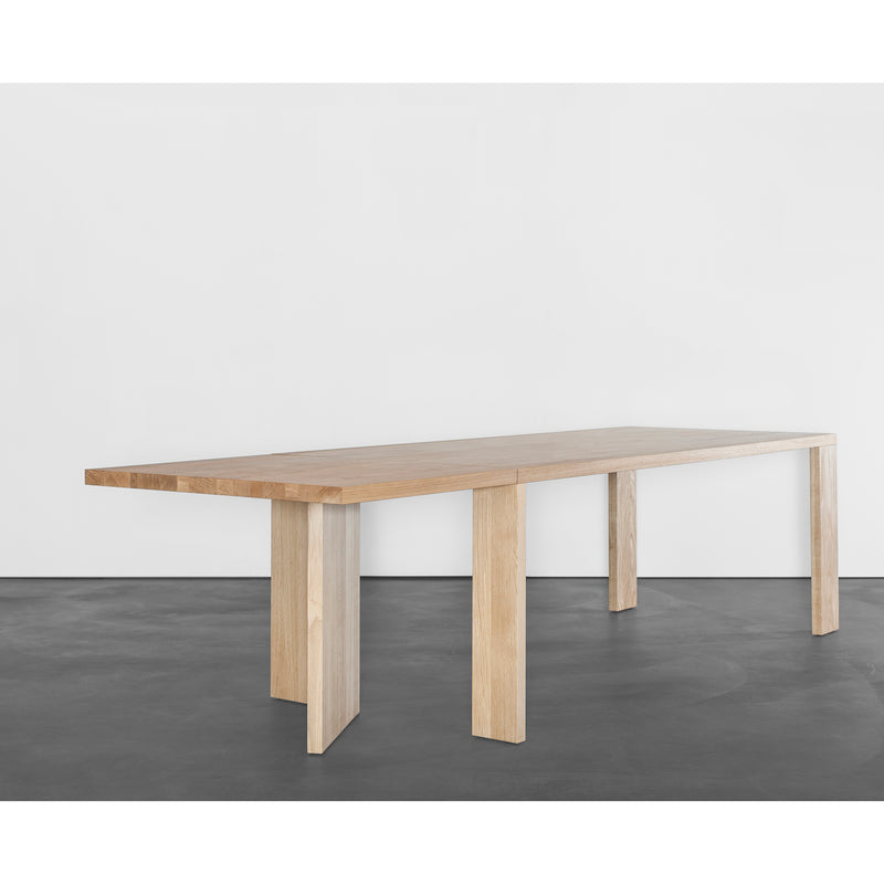 The Alphabet Folding Table by Sanktjohanser features a minimalist design with solid wood finishes, a rectangular top, and six sturdy legs. It stands out against a plain white background on a dark grey floor. Custom sizes are available to fit any dining space needs.