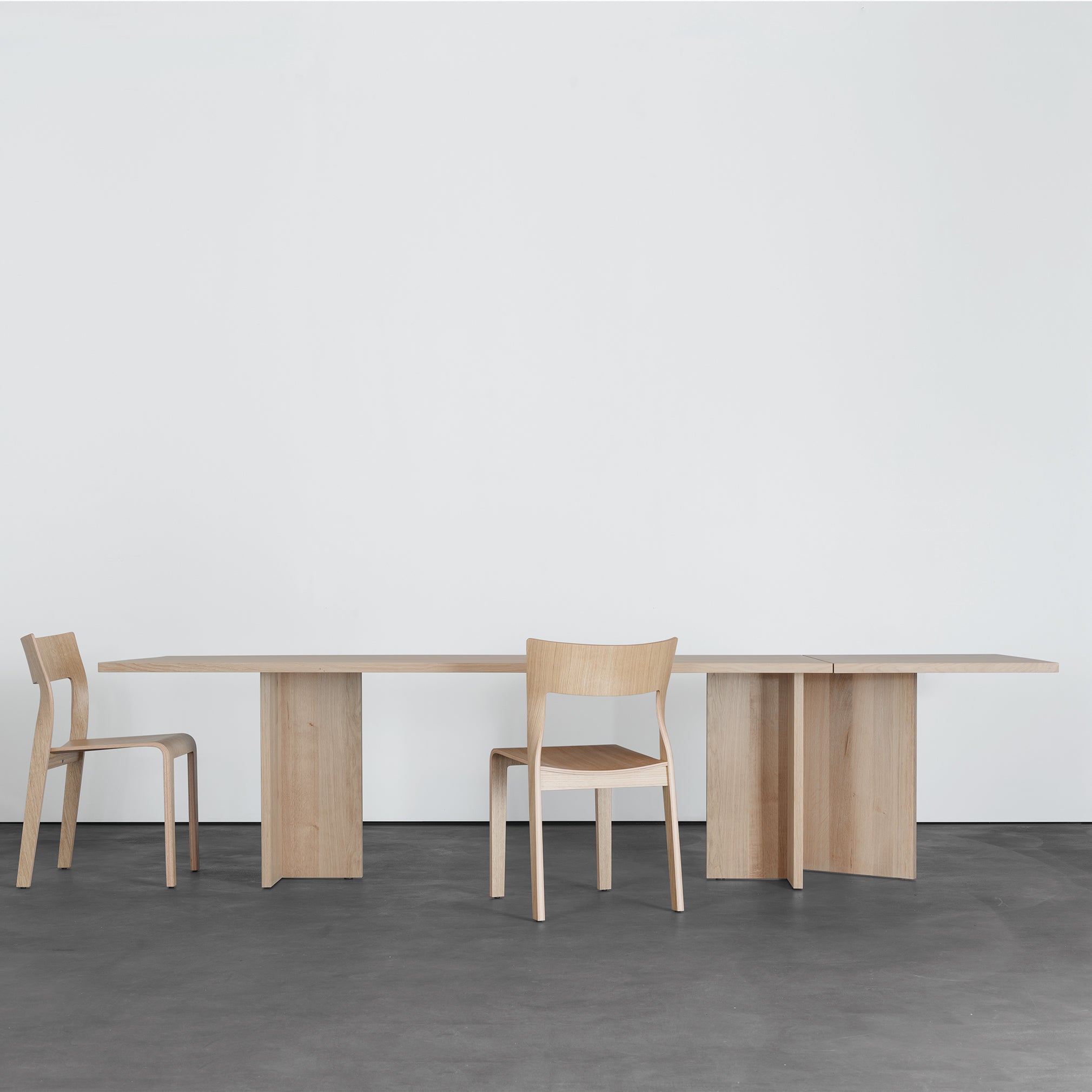 A minimalist room showcases a Sanktjohanser Alphabet Folding Table with solid wood finishes, paired with matching wooden chairs on a smooth dark floor. A plain white wall in the background enhances its sleek and modern look.
