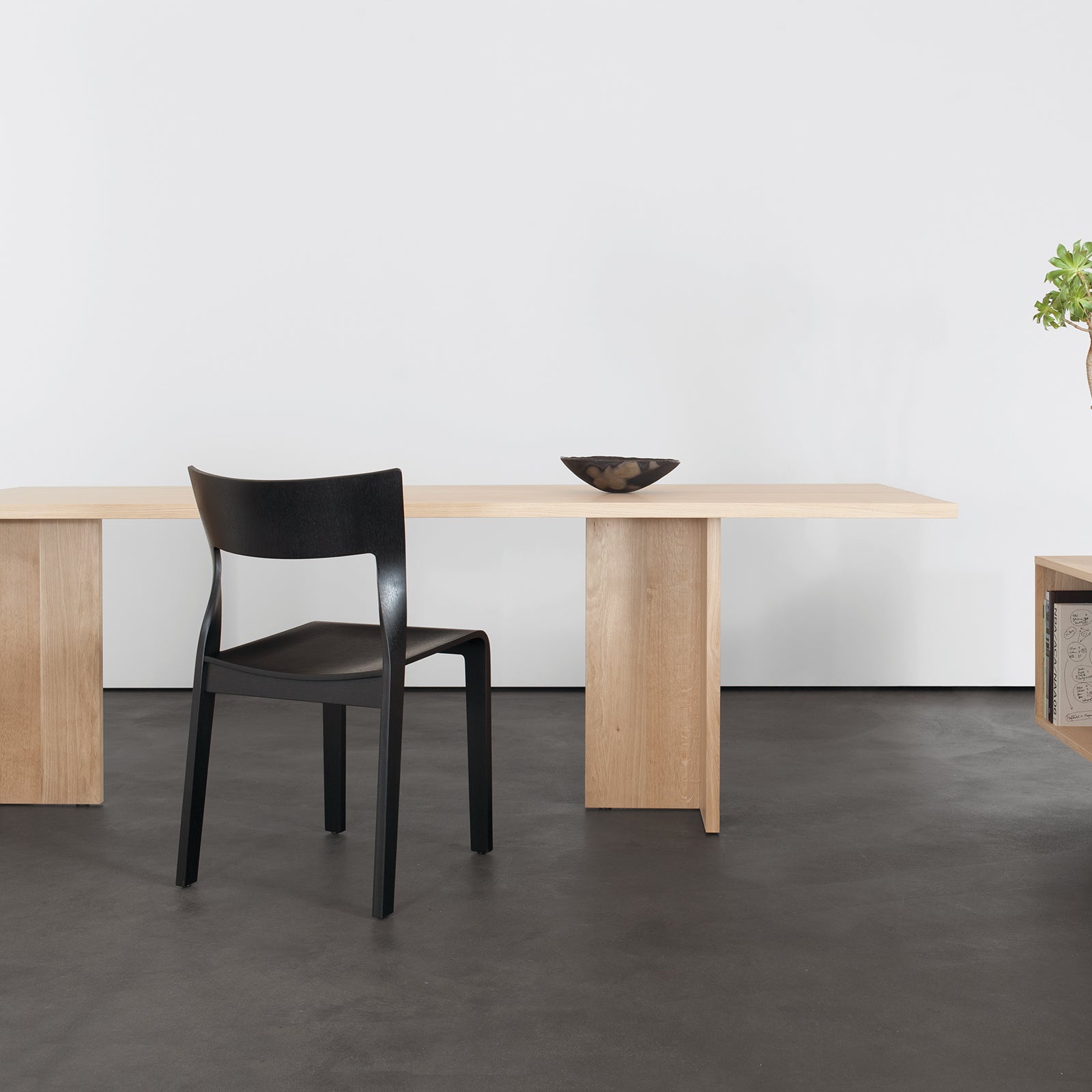 A minimalist room showcases a Sanktjohanser Alphabet Folding Table with solid wood finishes and a sleek black chair. A small decorative bowl graces the table, while a wooden shelf holds books and a potted plant. The space boasts modern charm with light floors, white walls, and custom sizes for flexibility.