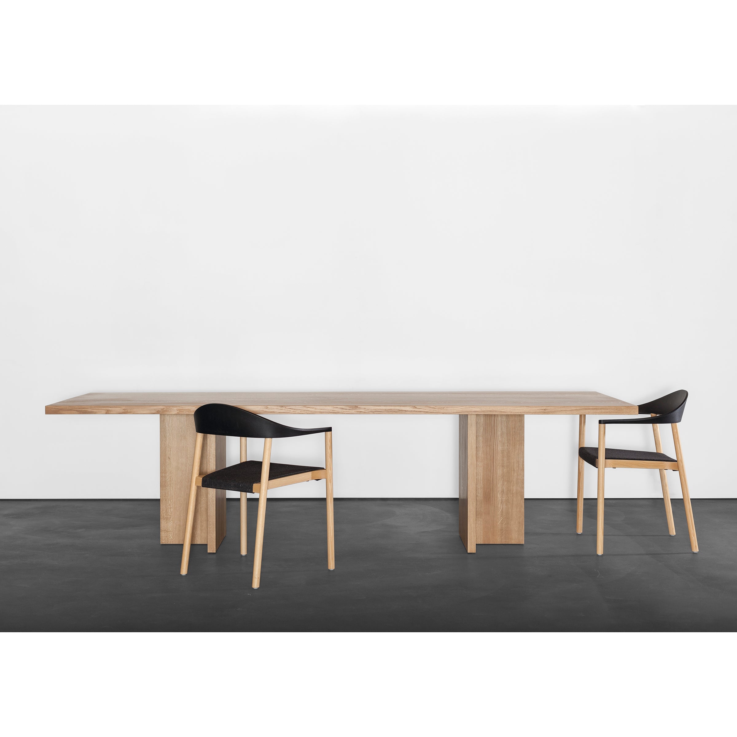 A minimalist room highlights a sleek Alphabet Table by Sanktjohanser with solid wood finishes and two black chairs, each featuring one of 26 leg designs. The tables light finish contrasts elegantly with the dark gray floor and plain white wall.