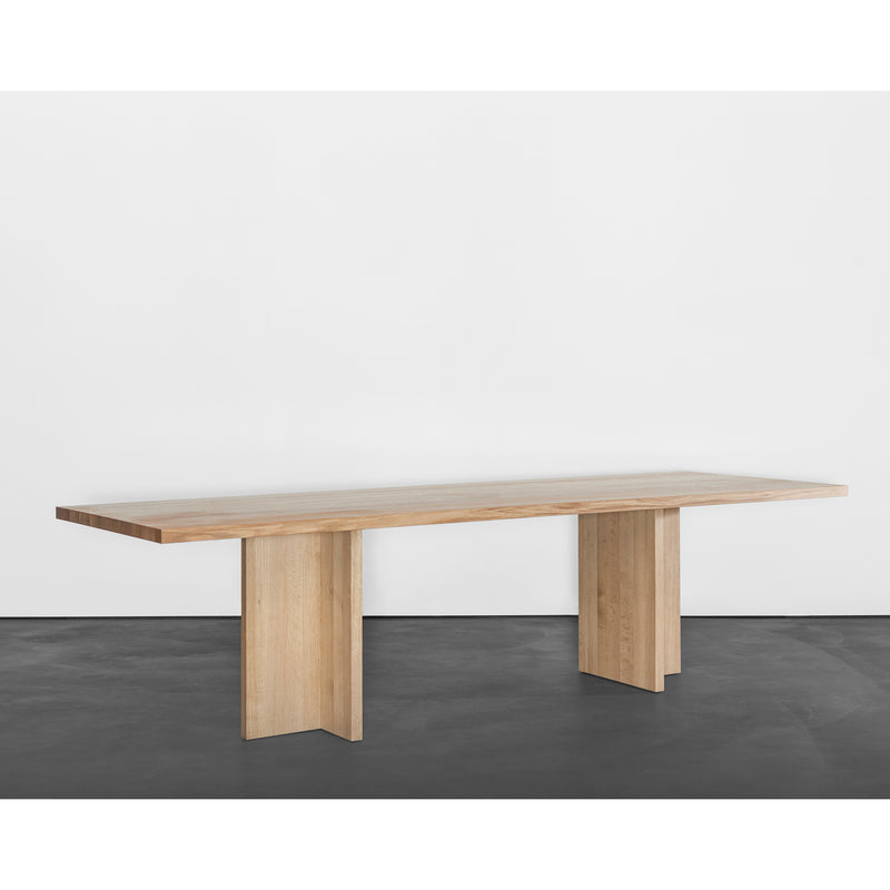 The Sanktjohanser Alphabet Table showcases a minimalist design with a flat rectangular surface and two sturdy vertical supports. With 26 leg designs available, its solid wood finish stands out against a white wall and dark floor.