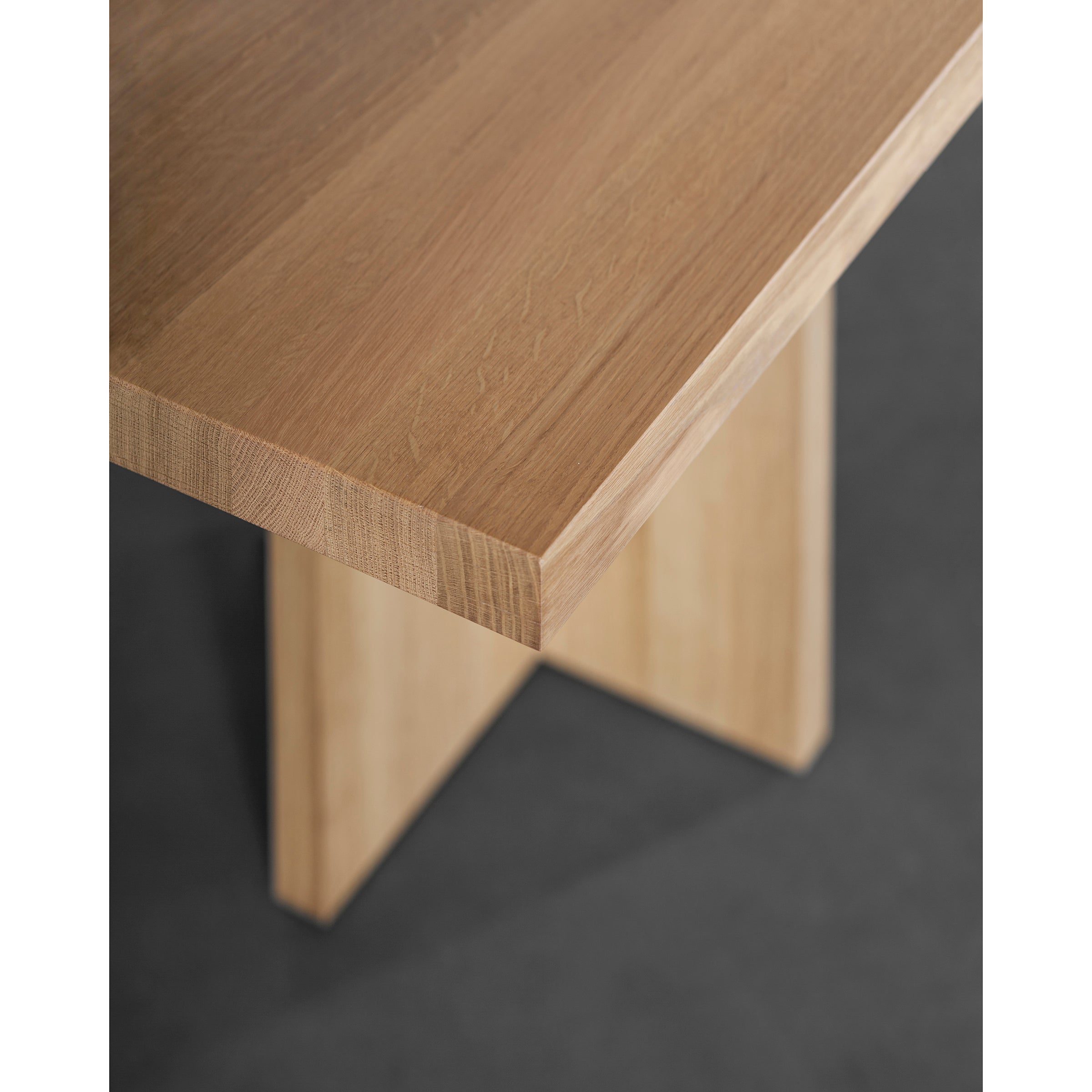 Close-up of the Alphabet Table by Sanktjohanser, showcasing a wooden corner with a natural finish. It features a thick, smooth top and sturdy X-shaped legs. The dark background enhances the solid woods texture and grains.
