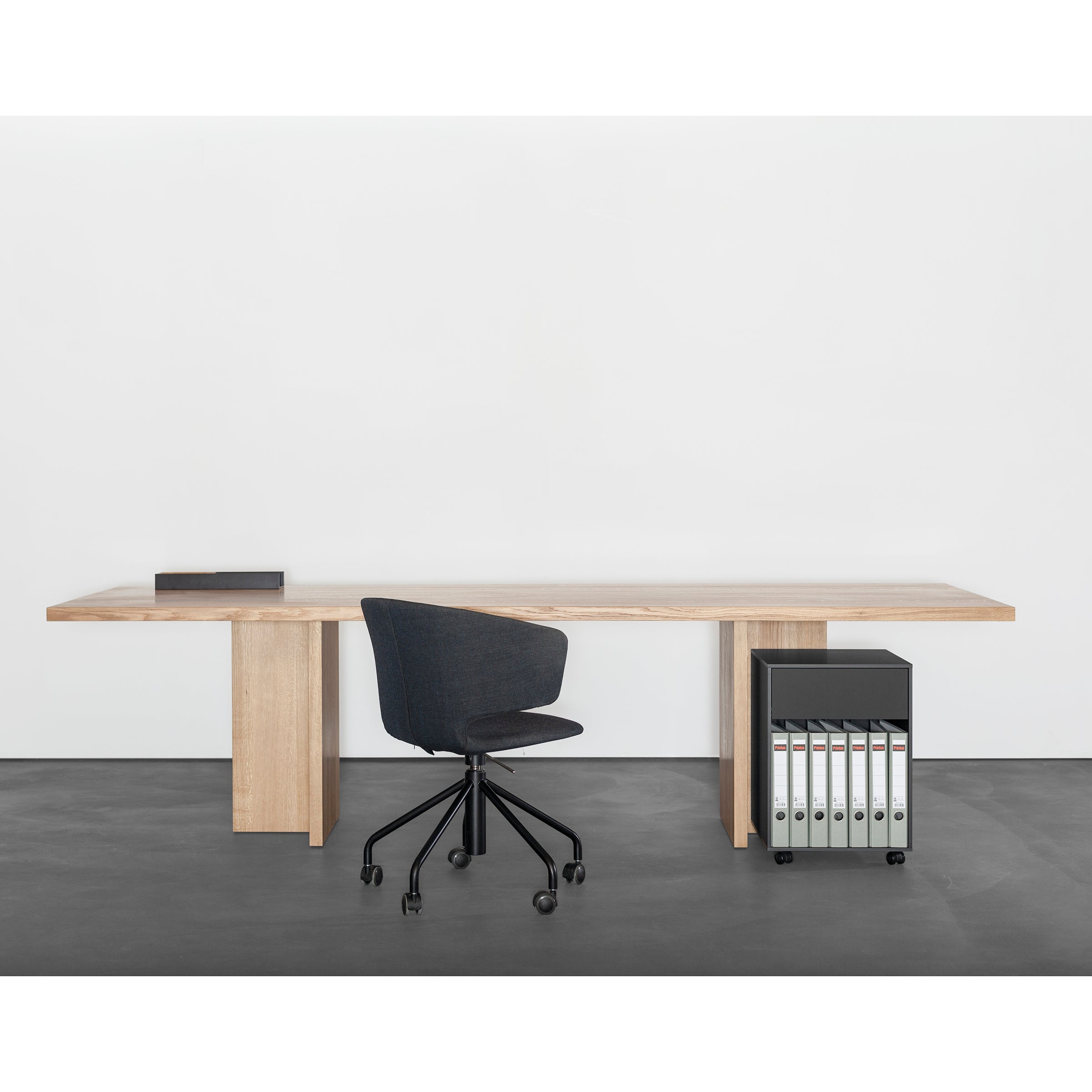A minimalist office setup features a Sanktjohanser Alphabet Table, black office chair, binders, and small black box on the desk. The plain white wall and dark gray floor enhance the sleek elegance of this space.