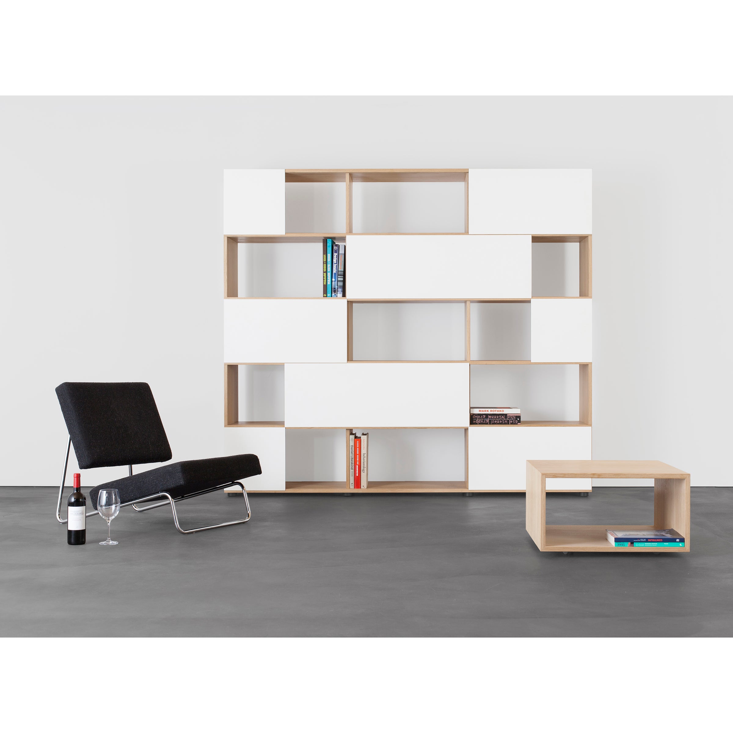 Modern living room with a black lounge chair, small wooden coffee table, and Sanktjohansers Analog multipurpose shelf in white and wood. The shelf holds colorful books. A wine bottle and glass are by the chair on sleek flooring beneath elegant sliding doors.