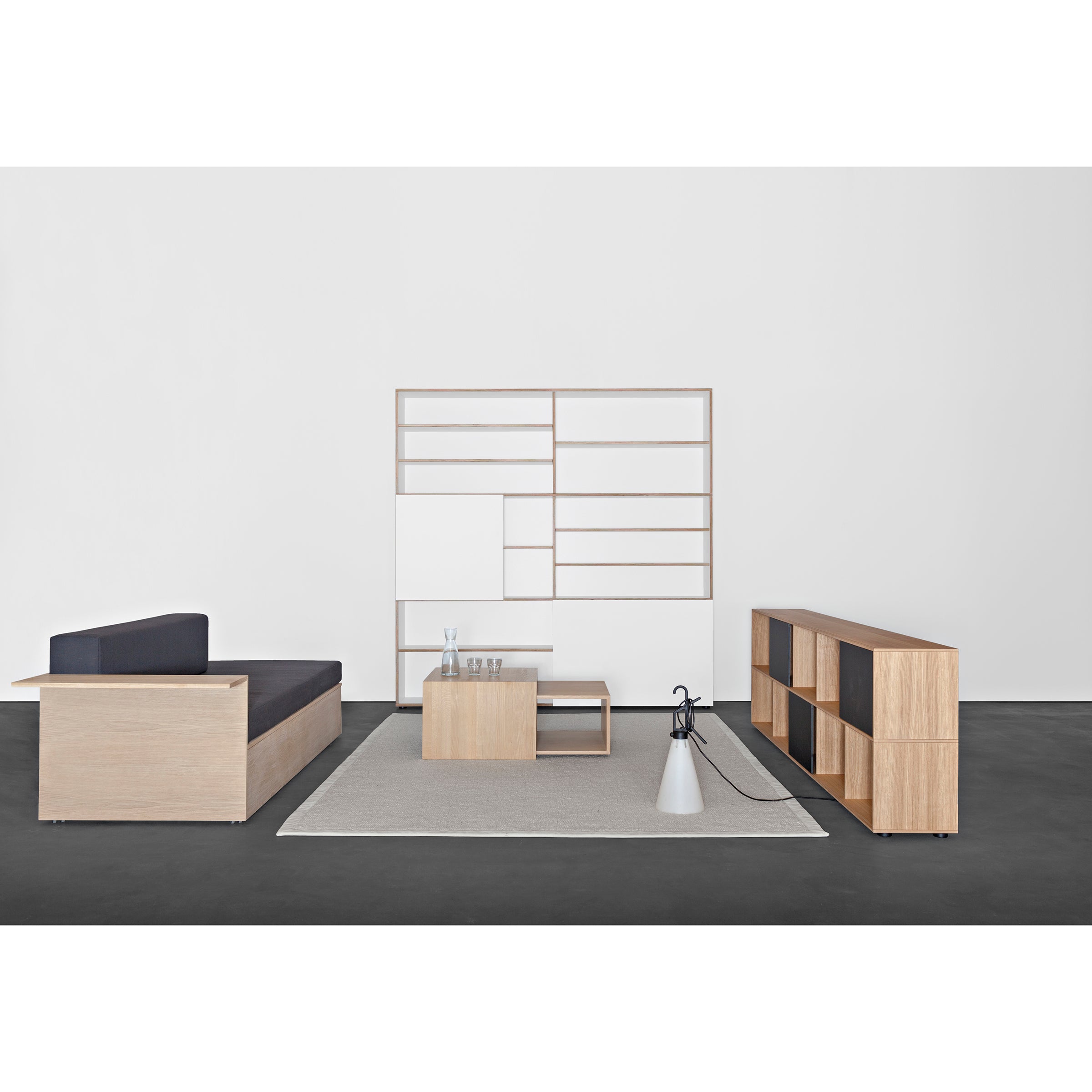 The minimalist living room features Sanktjohansers Analog multipurpose light solid wood furniture, including a low sofa and modular shelf unit. A gray rug enhances the dark floor, complemented by a mounted white panel. A modern floor lamp and decorative items add elegance to this harmonious setting.