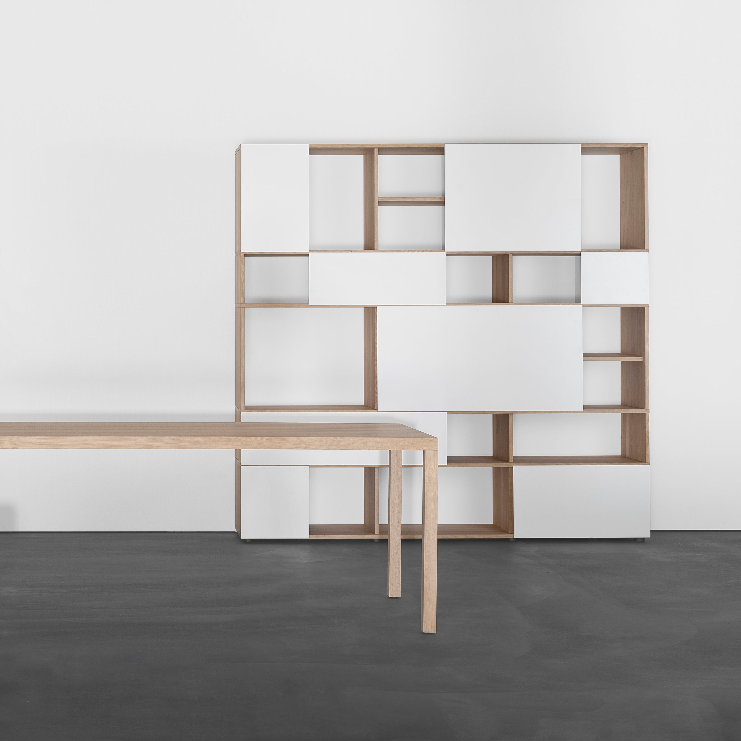 A minimalist room showcases a light wood table on a dark floor beside the sleek Analog multipurpose modular shelf by Sanktjohanser. The shelf features asymmetrical open wood shelves and closed white compartments on a plain white wall, with sliding doors enhancing the modern aesthetic.