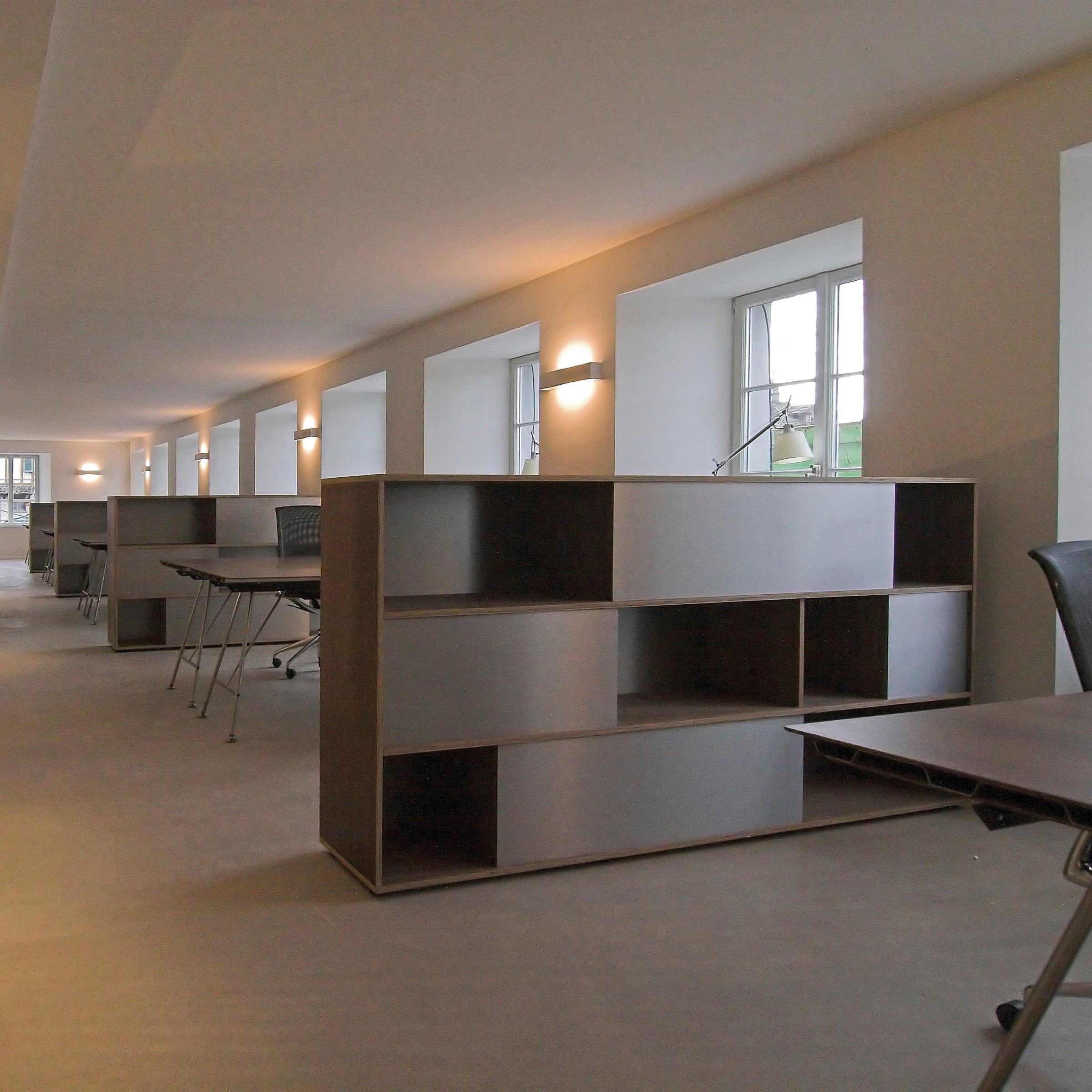 The well-lit modern office features Analog multipurpose desks and chairs from Sanktjohanser. Large windows and wall-mounted lights illuminate the space, while sliding doors provide privacy. Tall modular shelves divide the room, enhancing the clean, minimalistic decor.