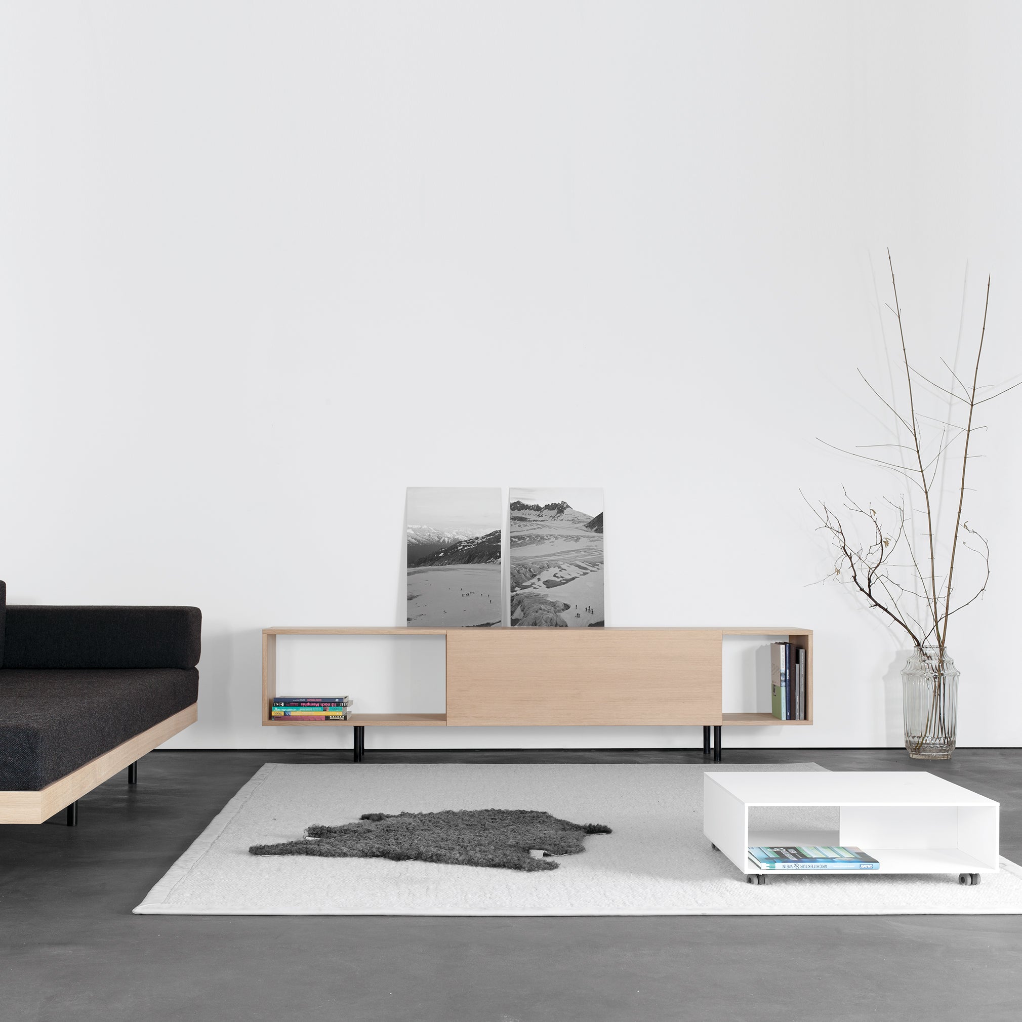 A minimalist living room features the Sanktjohanser Analog multipurpose wooden sideboard with two black and white photos, a bare branch in a vase, and a gray rug on light carpet. A low, white coffee table and dark sofa enhance the scene, illuminated by sliding doors that allow natural light.
