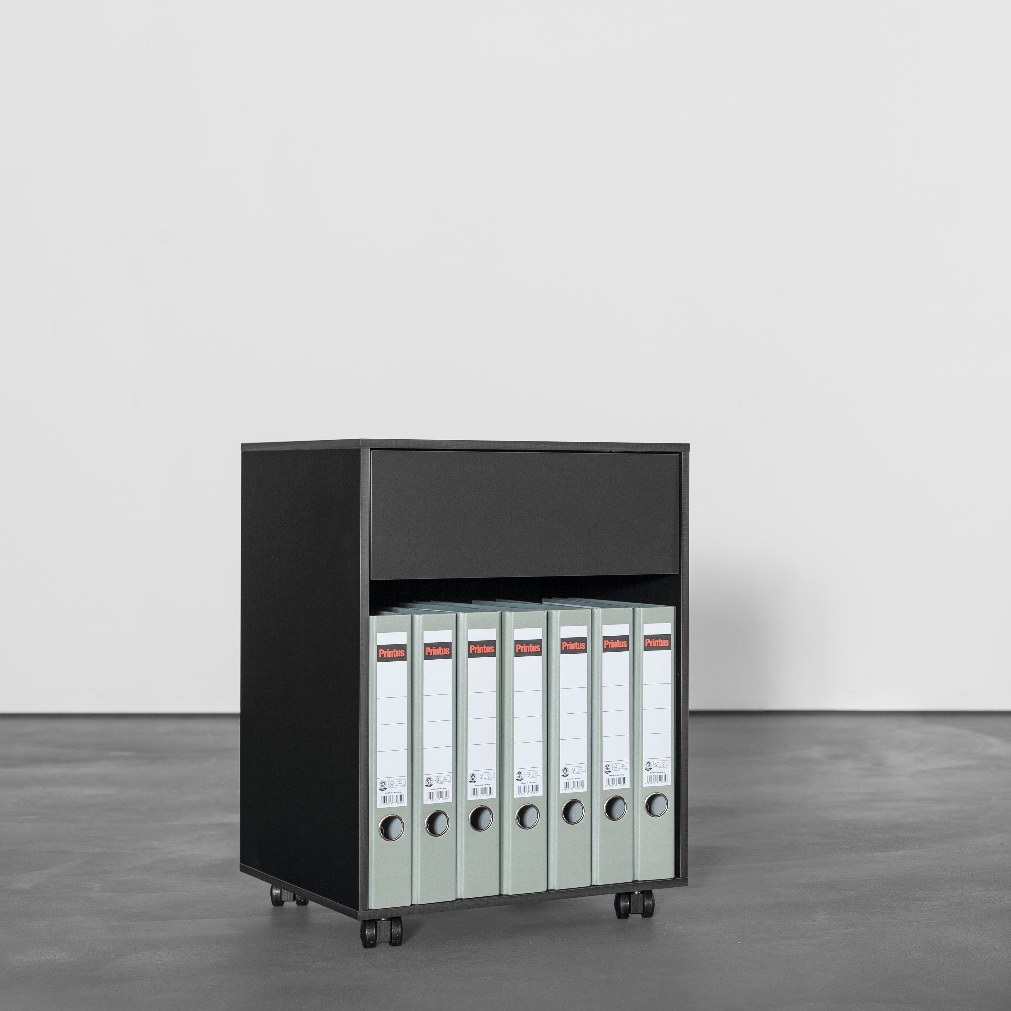 The black Sanktjohanser Annex Container on wheels is a sleek workplace organizer with an open shelf holding six green binders, set against a plain grey backdrop for modern practicality.