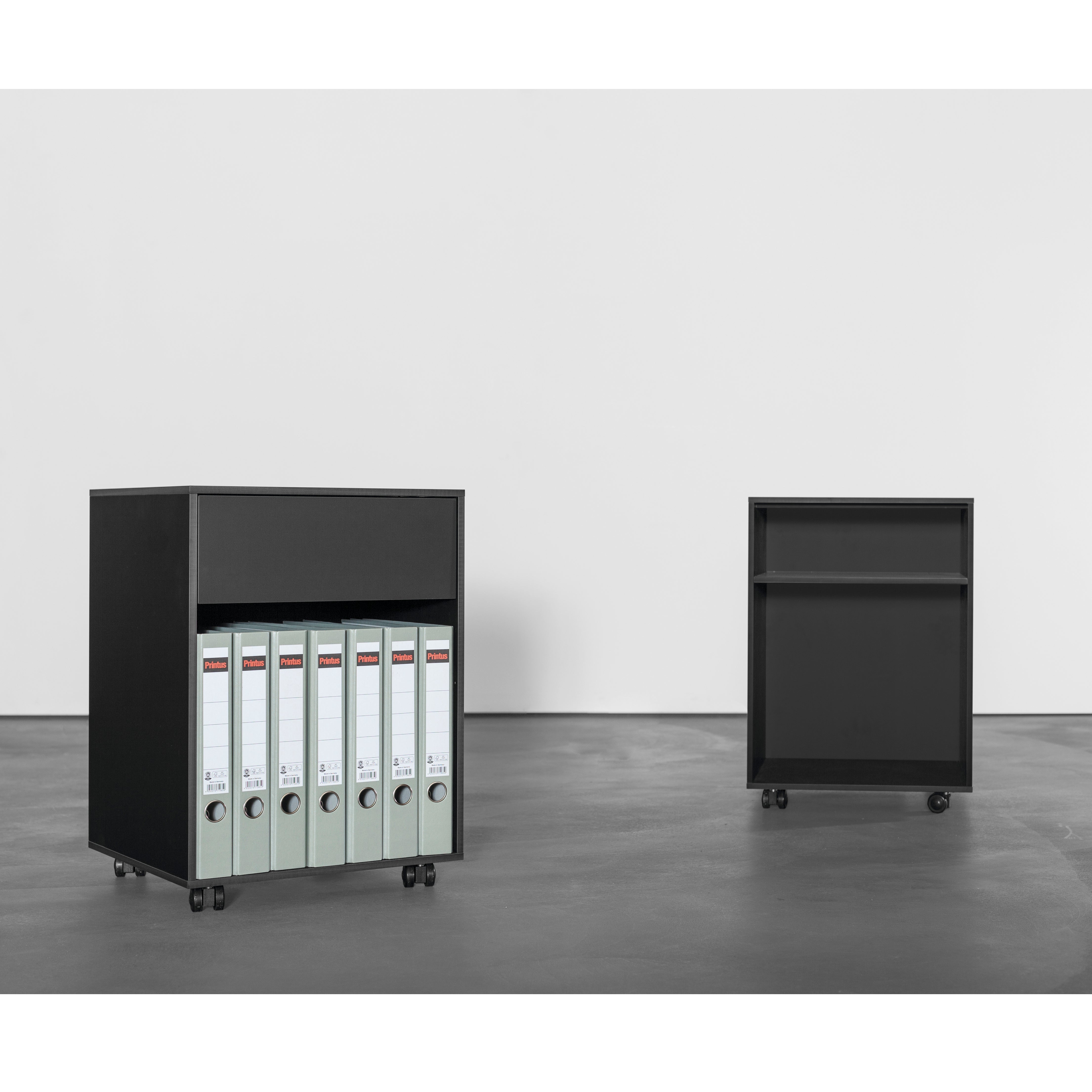 Two black Sanktjohanser Annex Containers, both on wheels, rest on a dark floor against a light wall. The left unit resembles an organizer with six binders inside, while the right is empty with two shelves. Both have a minimalist design and are made from sleek HPL or multiplex materials.