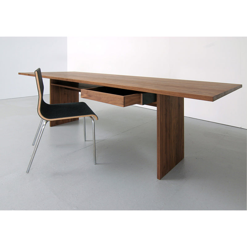 The stylish Areal Table by Sanktjohanser, featuring minimalist solid wood design and an open drawer, pairs perfectly with a modern chair with a black seat and metallic legs against a neutral background, exuding contemporary elegance.