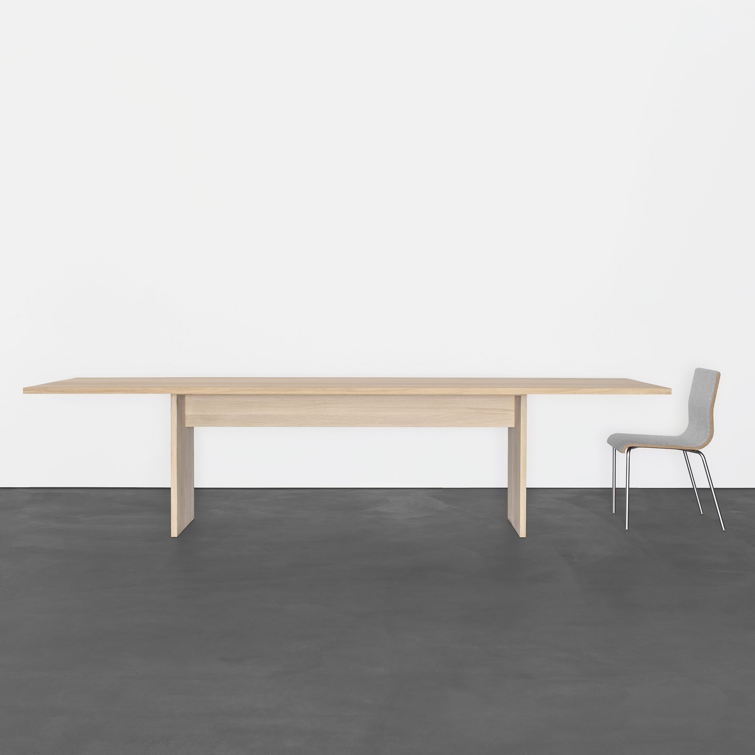 The minimalist room features a Sanktjohanser Areal Table with clean lines at its center, paired with a simple chair. The smooth dark gray floor and plain white wall enhance the modern atmosphere, while the tables solid wood construction adds elegance.