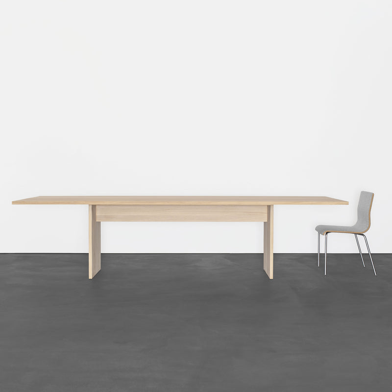 The minimalist room features a Sanktjohanser Areal Table with clean lines at its center, paired with a simple chair. The smooth dark gray floor and plain white wall enhance the modern atmosphere, while the tables solid wood construction adds elegance.
