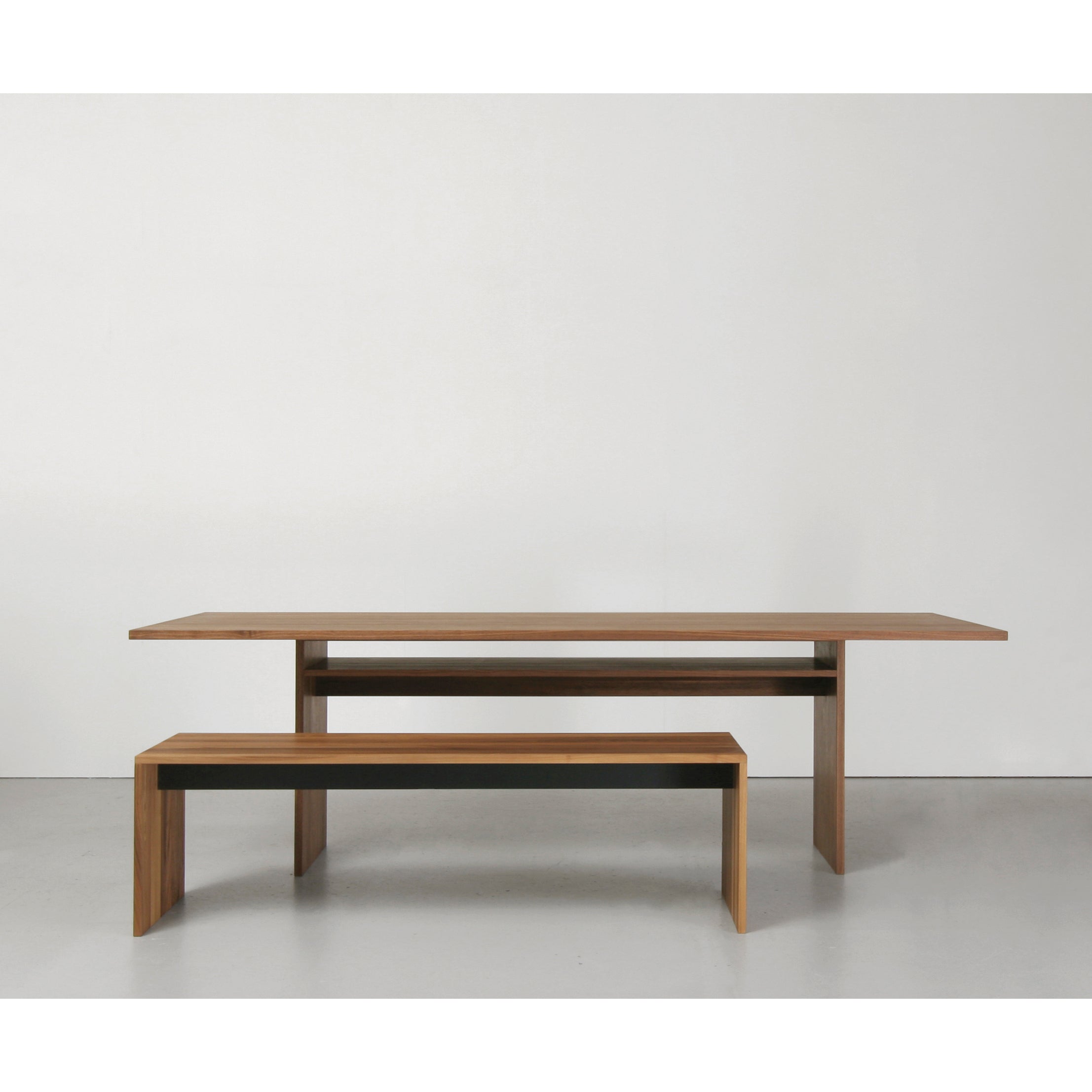 A modern wooden dining set against a plain white wall features the solid wood Areal Table by Sanktjohanser, accompanied by a matching bench. Both have clean lines and a minimalist design with black accents.