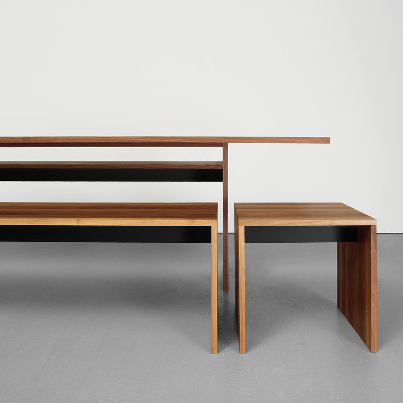 A minimalist furniture set featuring a long solid wood bench and the Sanktjohanser Areal Table, both with sharp, clean lines and elegant design. Set against a plain white wall on a smooth, light gray floor, this ensemble exudes modern sophistication.