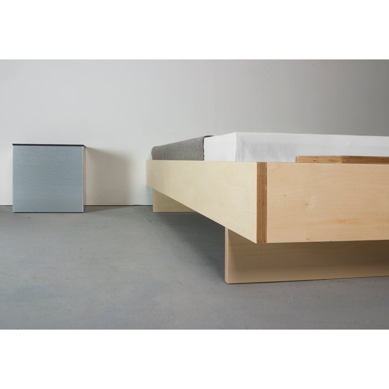 A minimalist bedroom features a Sanktjohanser Bezwei Bed with grey bedding. A small square nightstand with a light grey front stands against a white wall, offering sleek functionality. Customizable bed dimensions enhance the rooms simple, modern aesthetic.