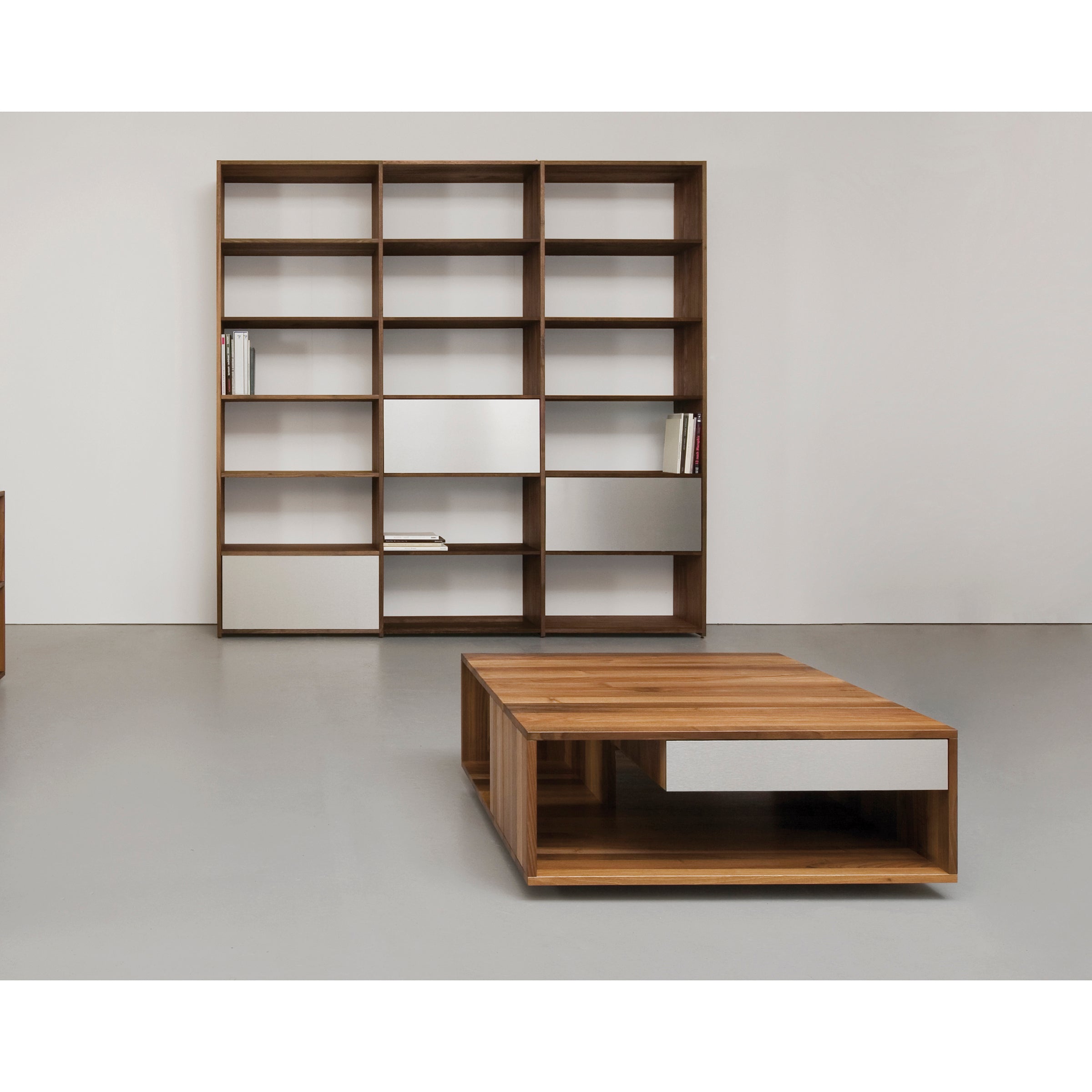 The minimalist room features a tall Sanktjohanser Dasregal shelf with open shelves and drawers. In the foreground, theres a low rectangular solid wood coffee table with open shelving. The walls and floor are light gray.