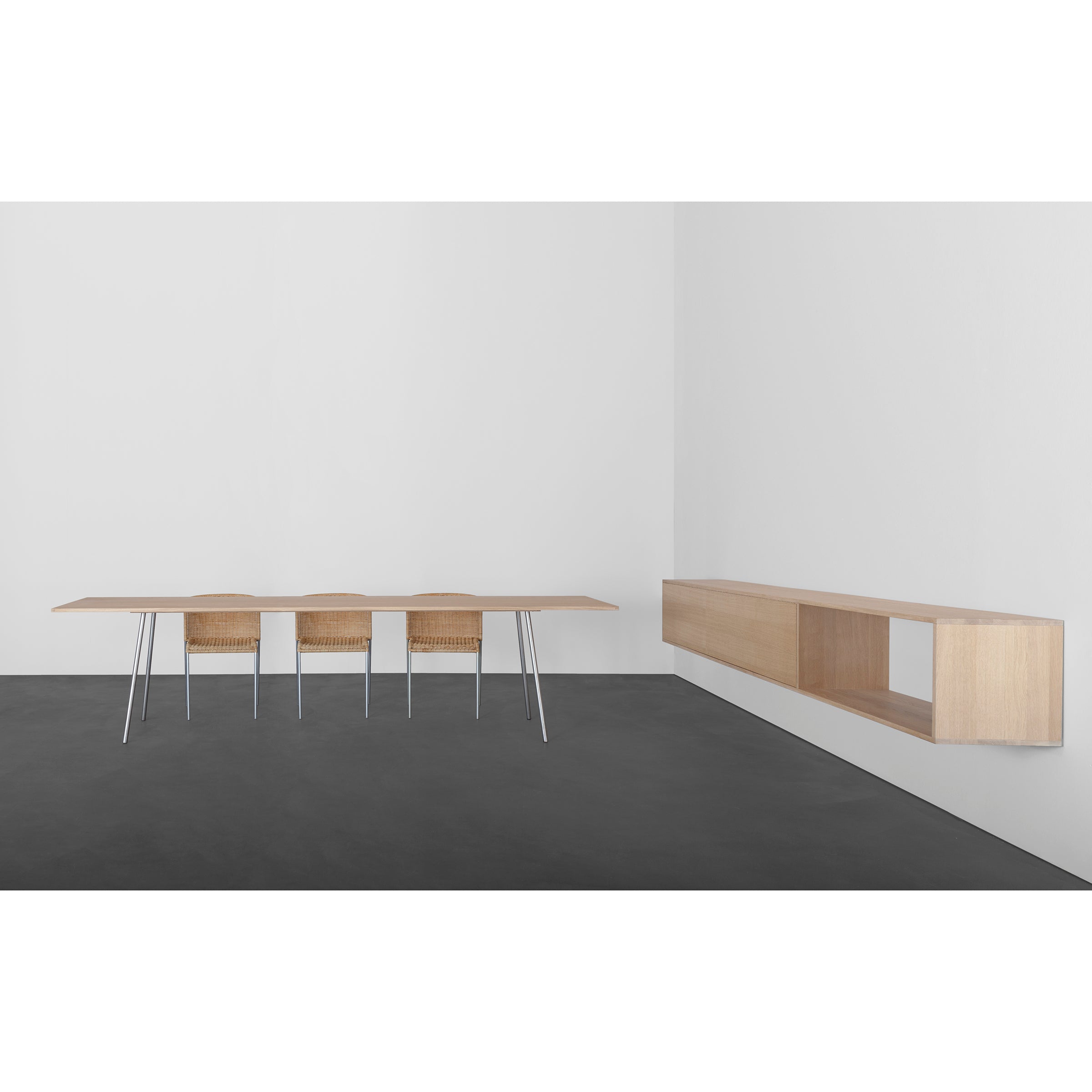 A minimalist room includes three solid wood chairs and a long table with slender metal legs against gray floors and white walls. The right wall is adorned with a functional Dasregal shelf by Sanktjohanser, featuring an open compartment for elegant storage.