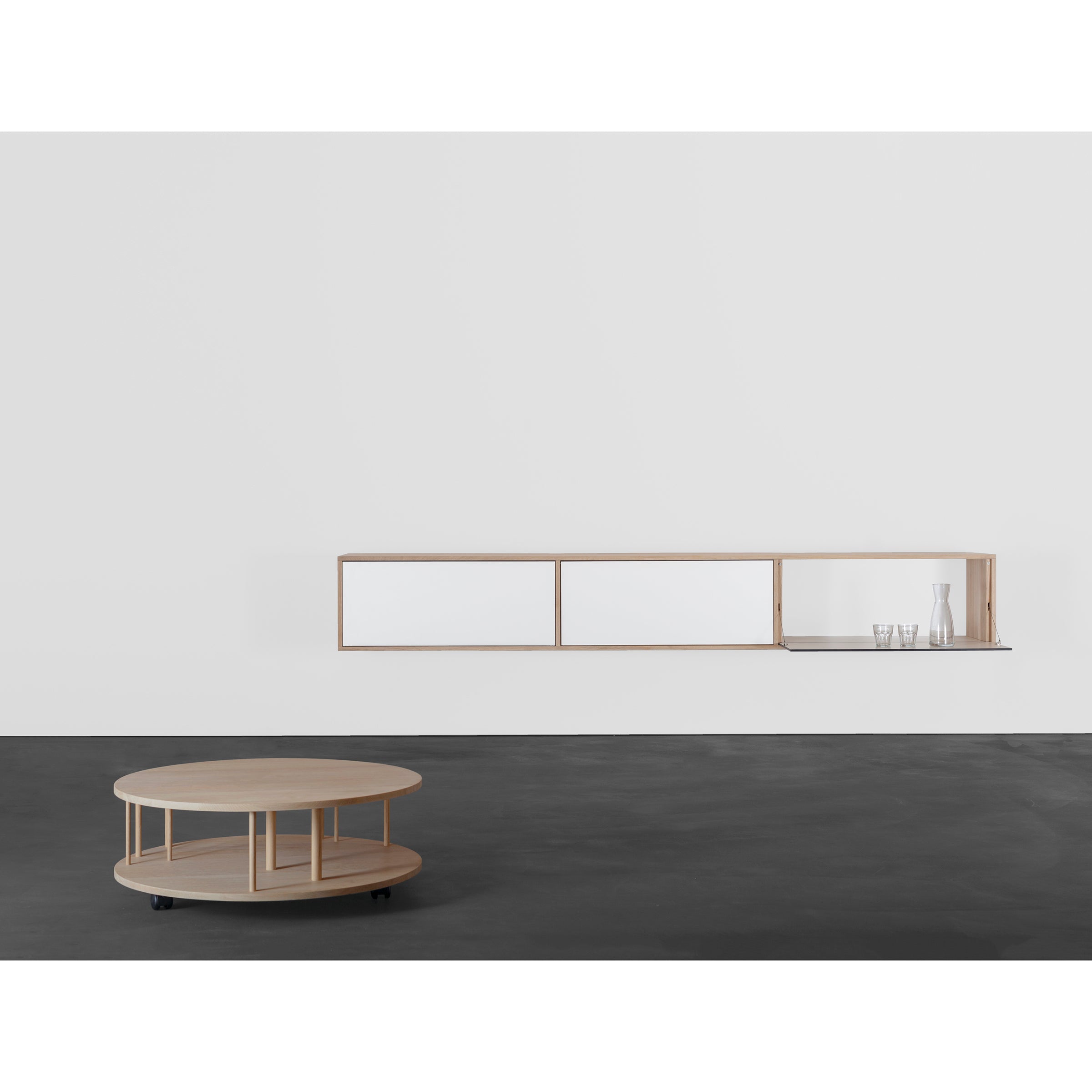 A minimalist room features the sleek wall-mounted Sanktjohanser Dasregal shelf system with closed cabinets. A round light wood coffee table with a wheeled lower shelf stands on the dark floor, while a carafe and glasses are elegantly placed on the Dasregal shelf.