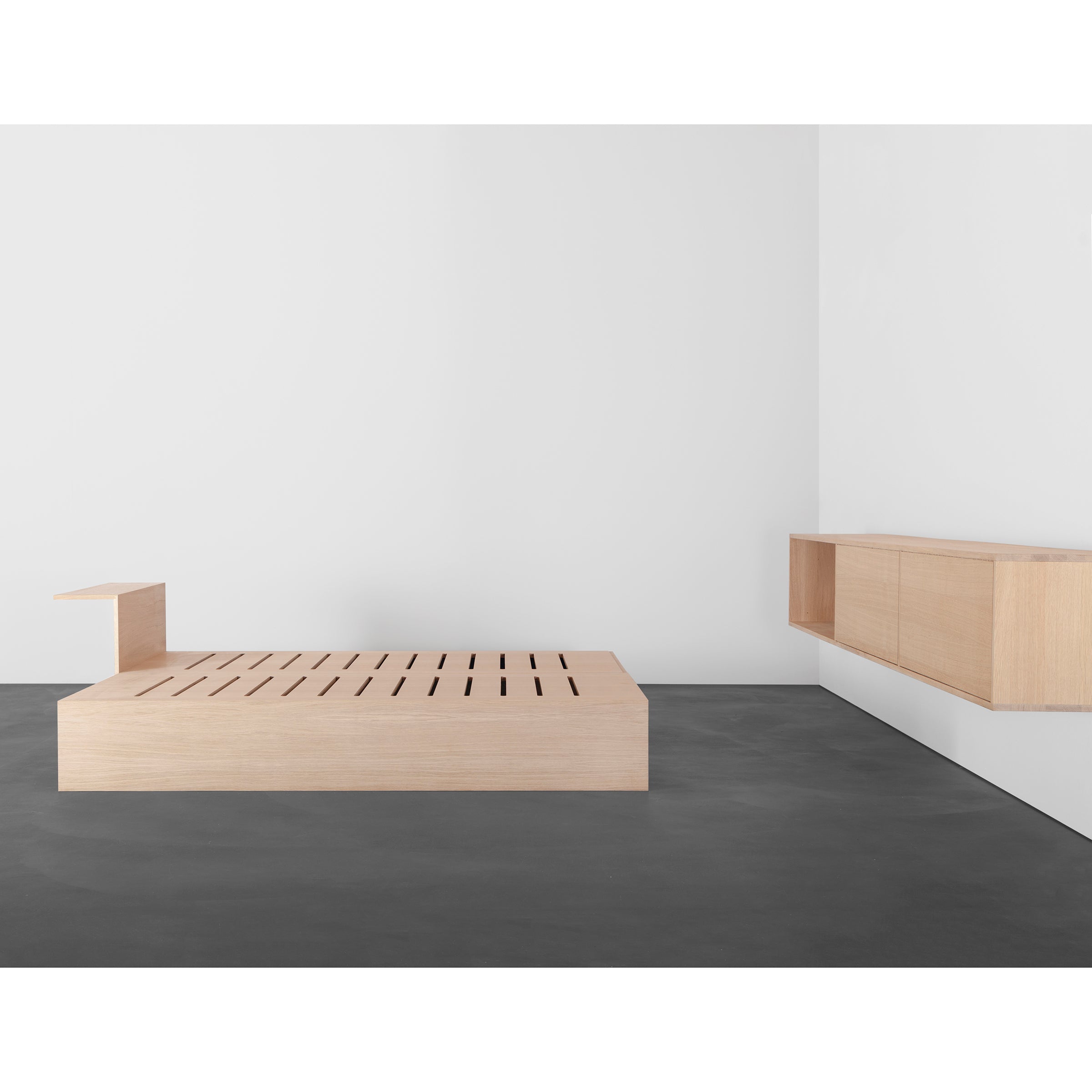 A minimalist room features a light wood platform bed with a slatted design and attached side table. A Dasregal shelf from Sanktjohanser, wall-mounted and customizable in matching wood, complements the floating cabinet on the white wall while the dark gray floor contrasts with the solid wood furniture.