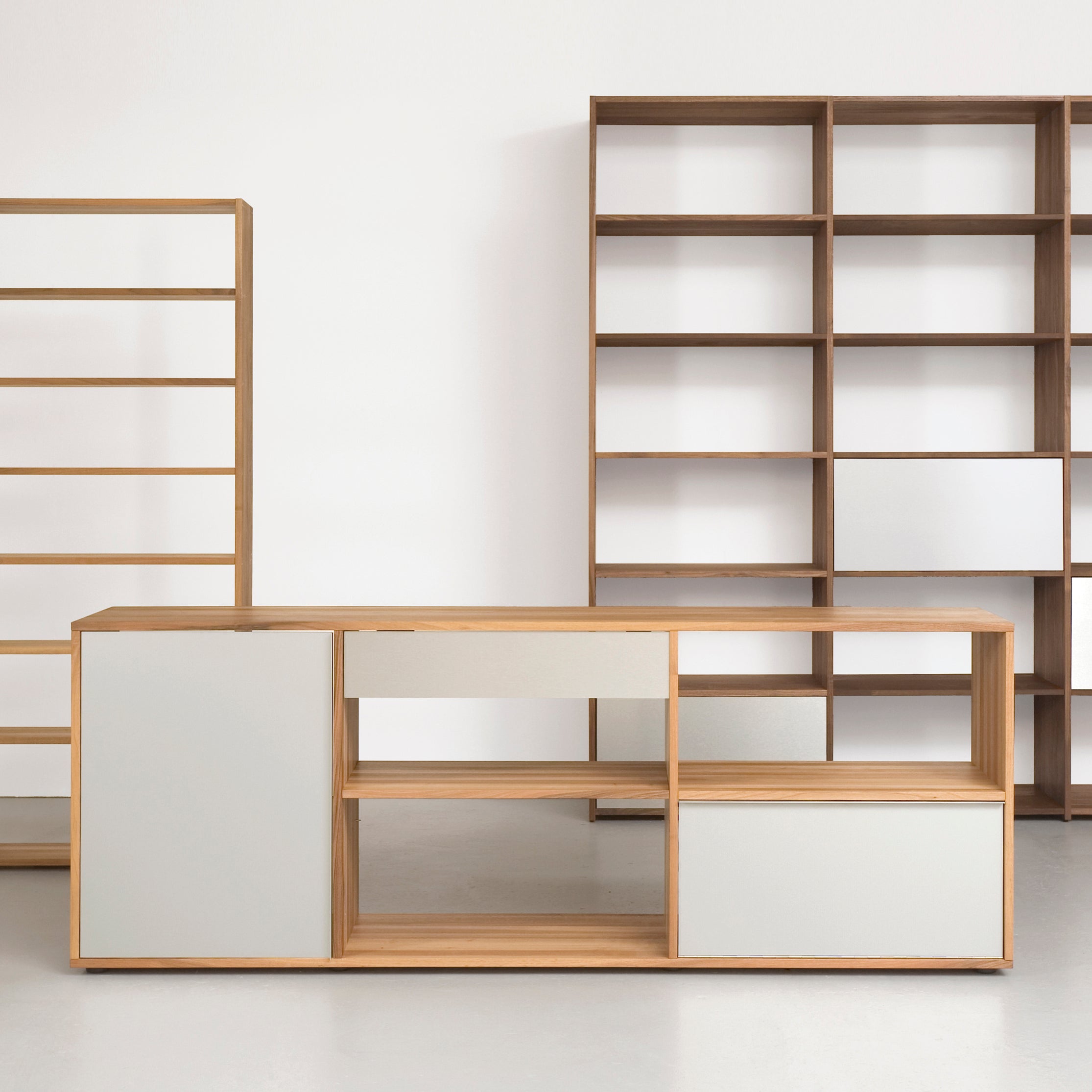 The Dasregal shelf by Sanktjohanser is a minimalist wooden furniture set with open shelving, closed cabinets, and a wall-mounted shelf. It features clean lines, a natural wood finish with white panels, and its neutral background enhances the modern aesthetic of these solid wood pieces.