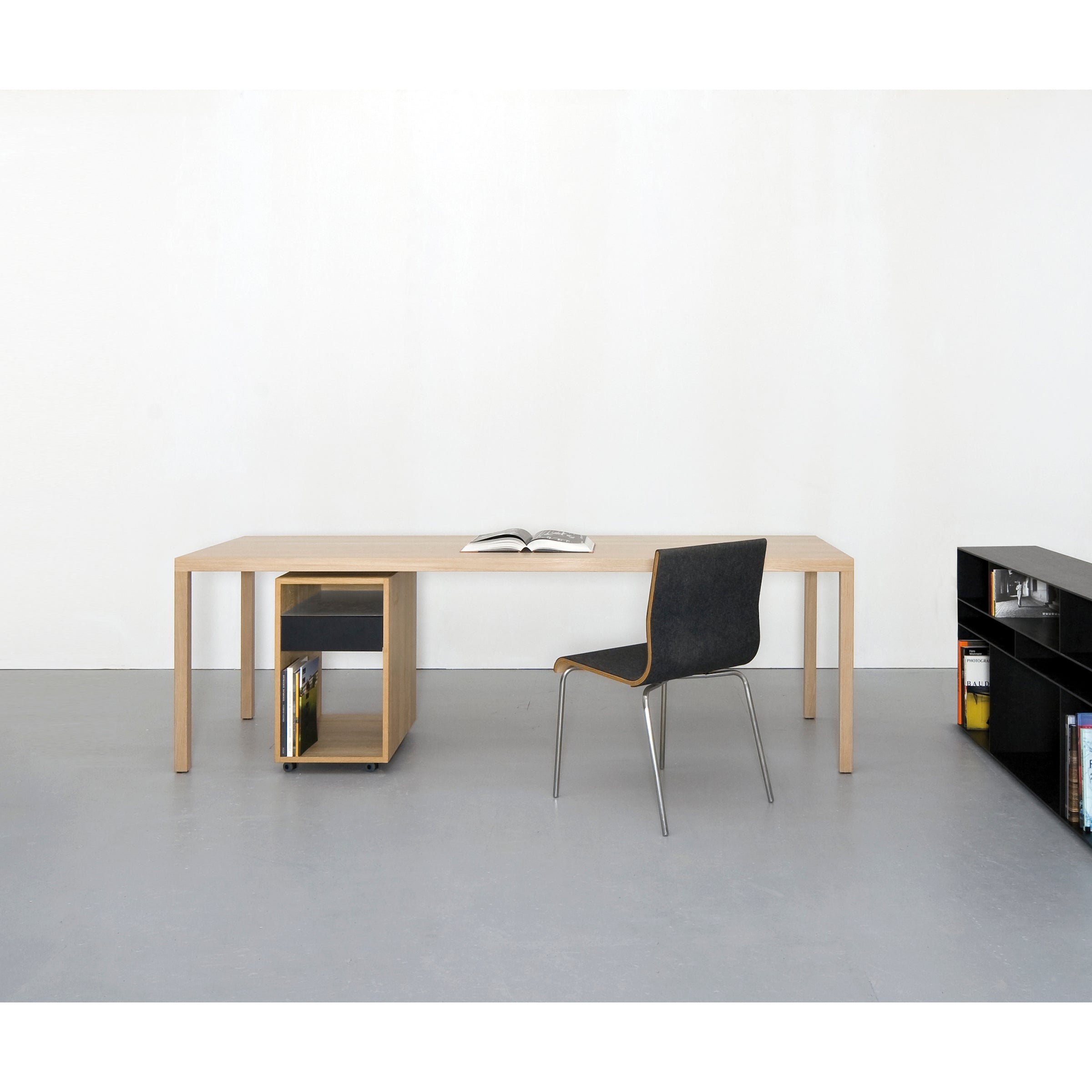 A minimalist room includes a light wooden table with an open book and a black chair. A small shelf serves as a side table, and a larger black shelving unit by Sanktjohanser, like the Depot X Container, is partially visible on the right.
