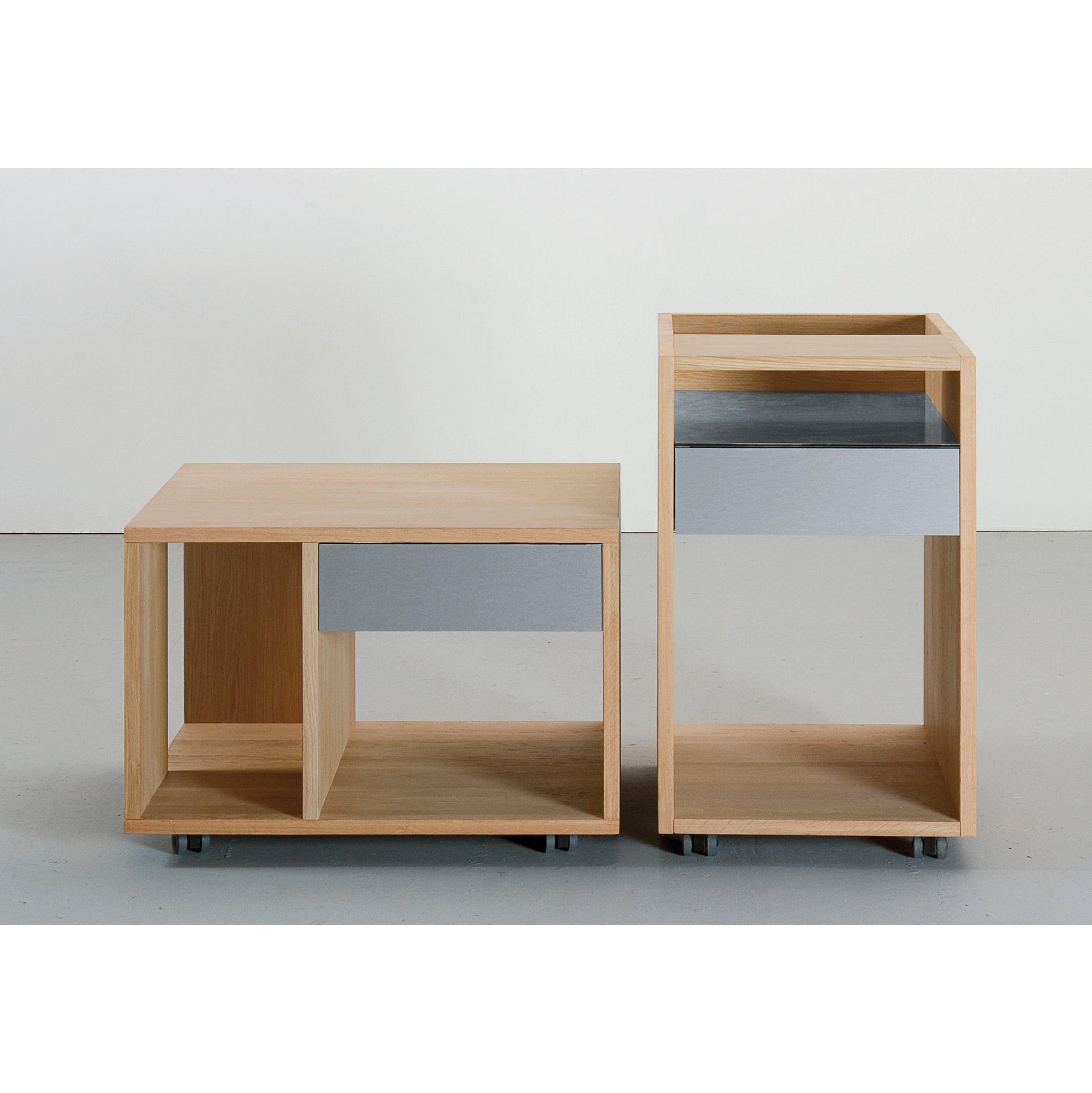 The Sanktjohanser Depot X Container set includes two modern wooden side tables with open shelving. One taller piece has a metallic drawer, both have wheels for mobility. The minimalist design showcases clean lines and a natural wood finish, ideal for storing essentials.