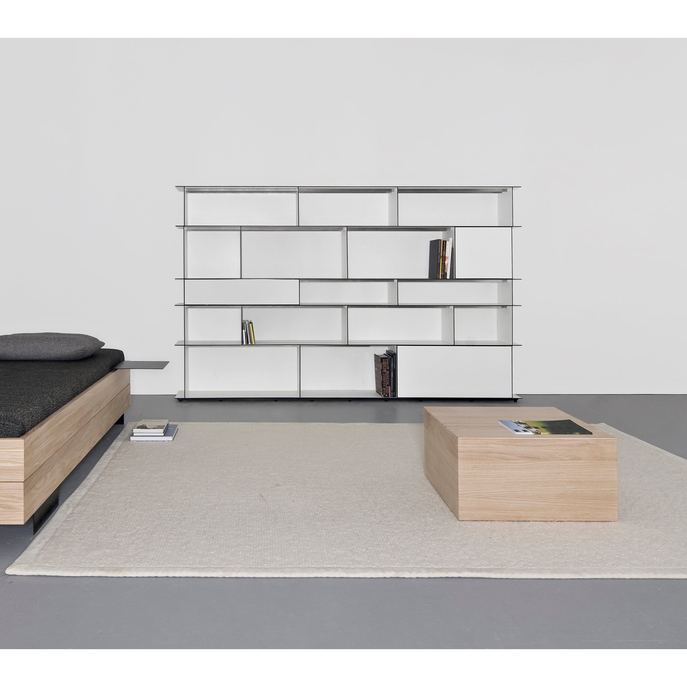 A minimalist room showcases Sanktjohansers Digital multipurpose shelf system; a modern, white geometric unit partially filled with books against a white wall. A gray cushioned wooden bench and a small wooden coffee table rest on a light gray rug.