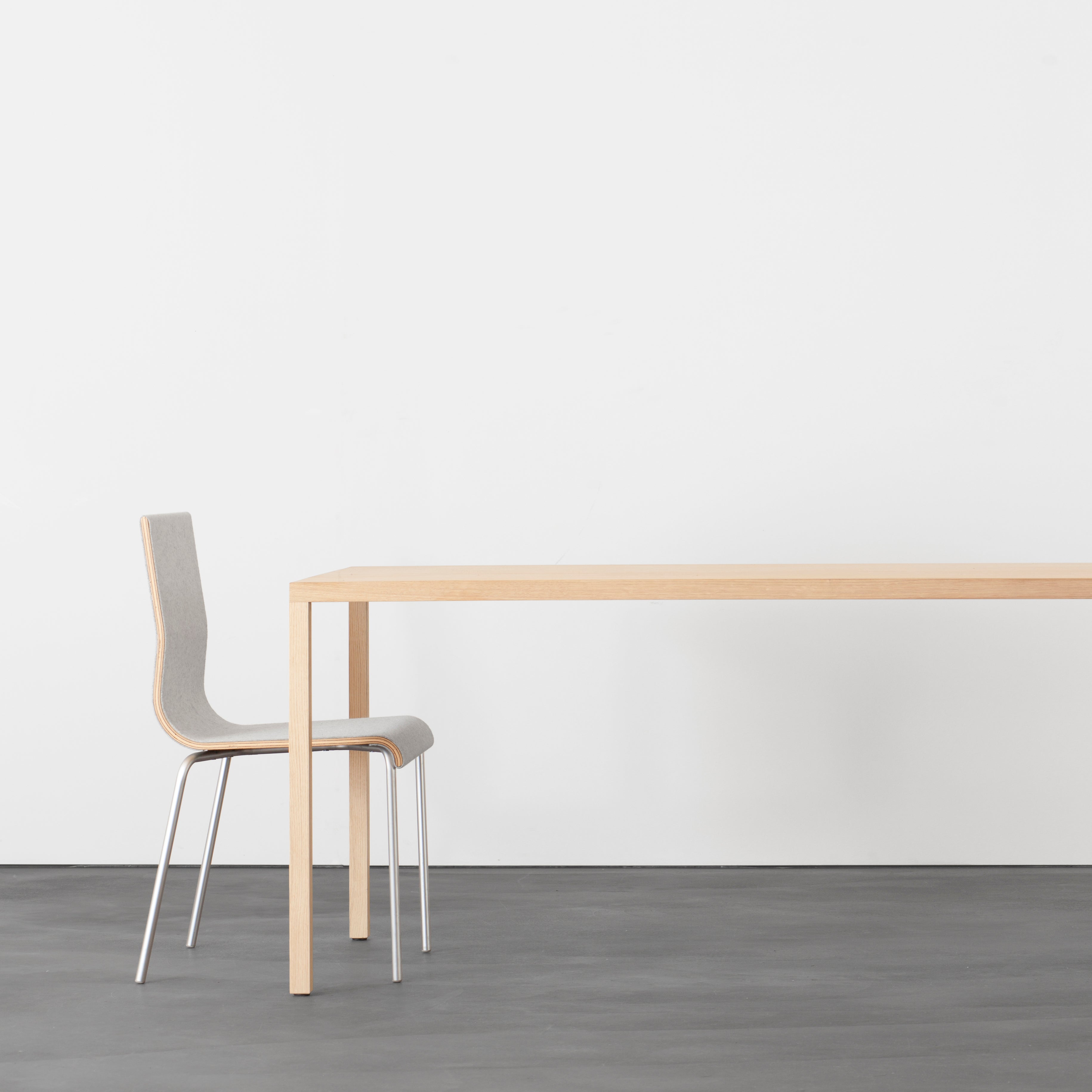 A minimalist room showcases a sleek Din36 Table by Sanktjohanser made from solid wood, paired with a gray chair on a dark floor. The plain white wall in the background accentuates the simplicity and modernity of this stylish space.