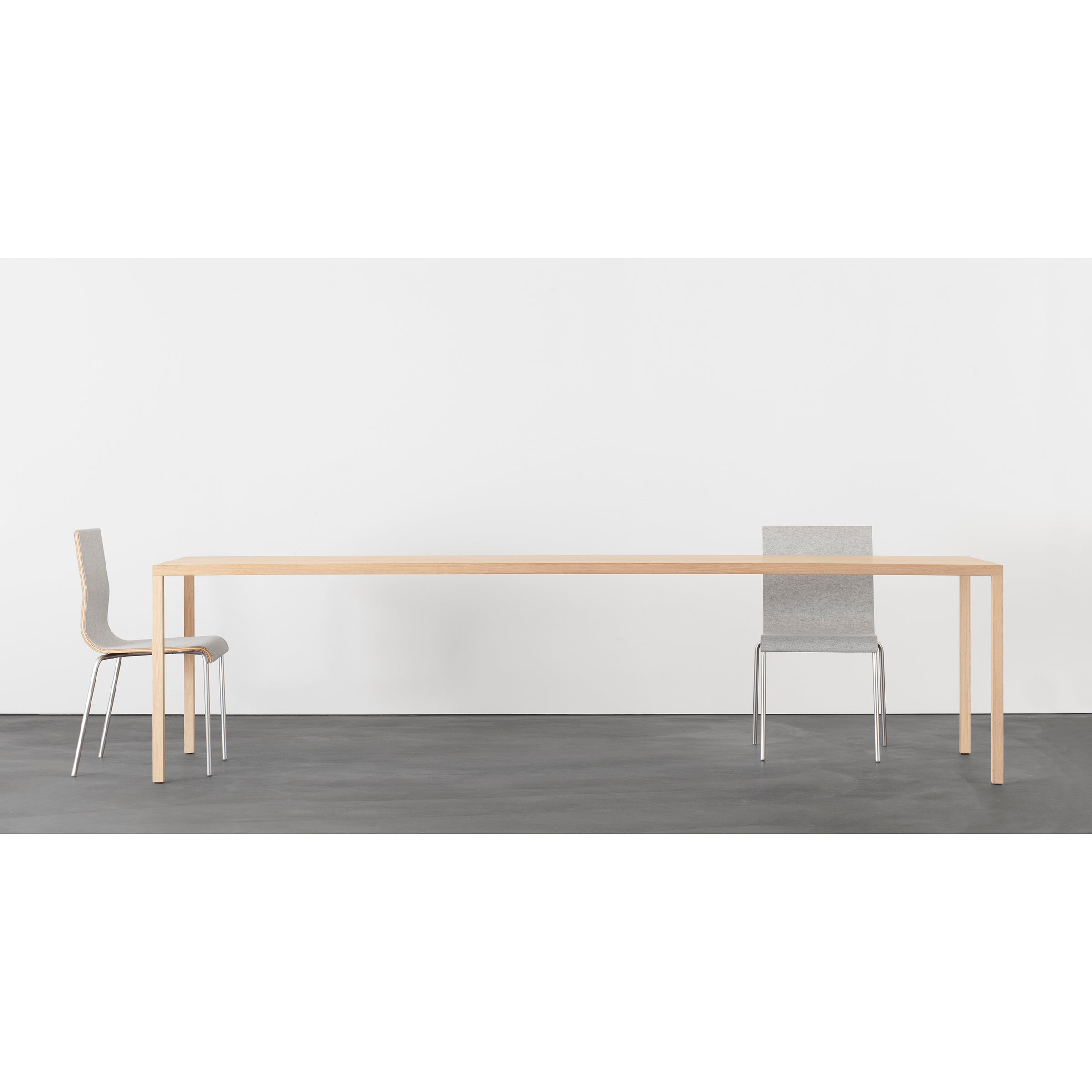 A minimalist setting with a long, light solid wood dining table by Sanktjohanser, reminiscent of the Din36 Table. Two sleek chairs with thin steel legs flank the table on either side. The plain white background contrasts with the dark grey floor.