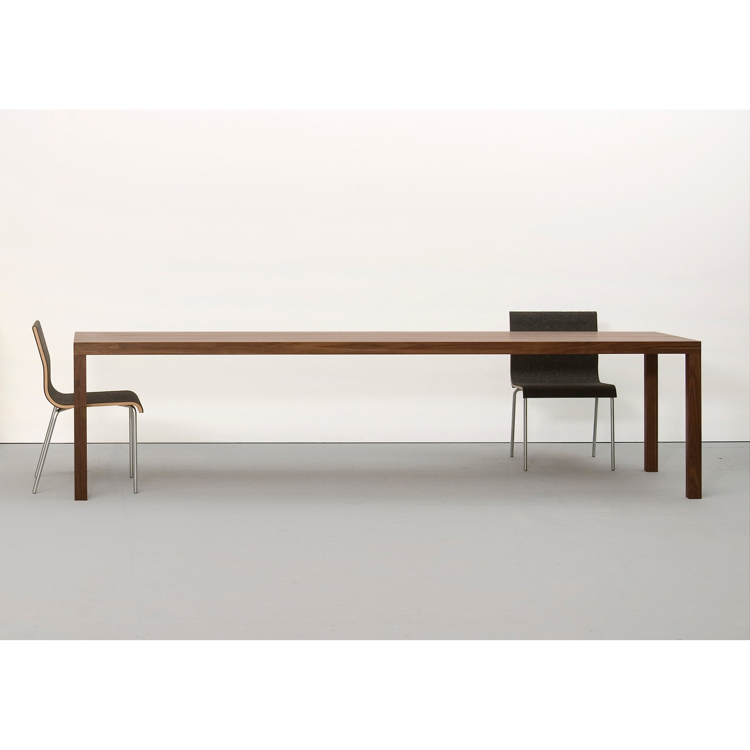 In a minimalist scene, the Sanktjohanser Dinavier Table, known for its detachable legs and solid wood design, is styled with two chairs at each end. A white background and floor enhance the simplicity of this long, rectangular table.