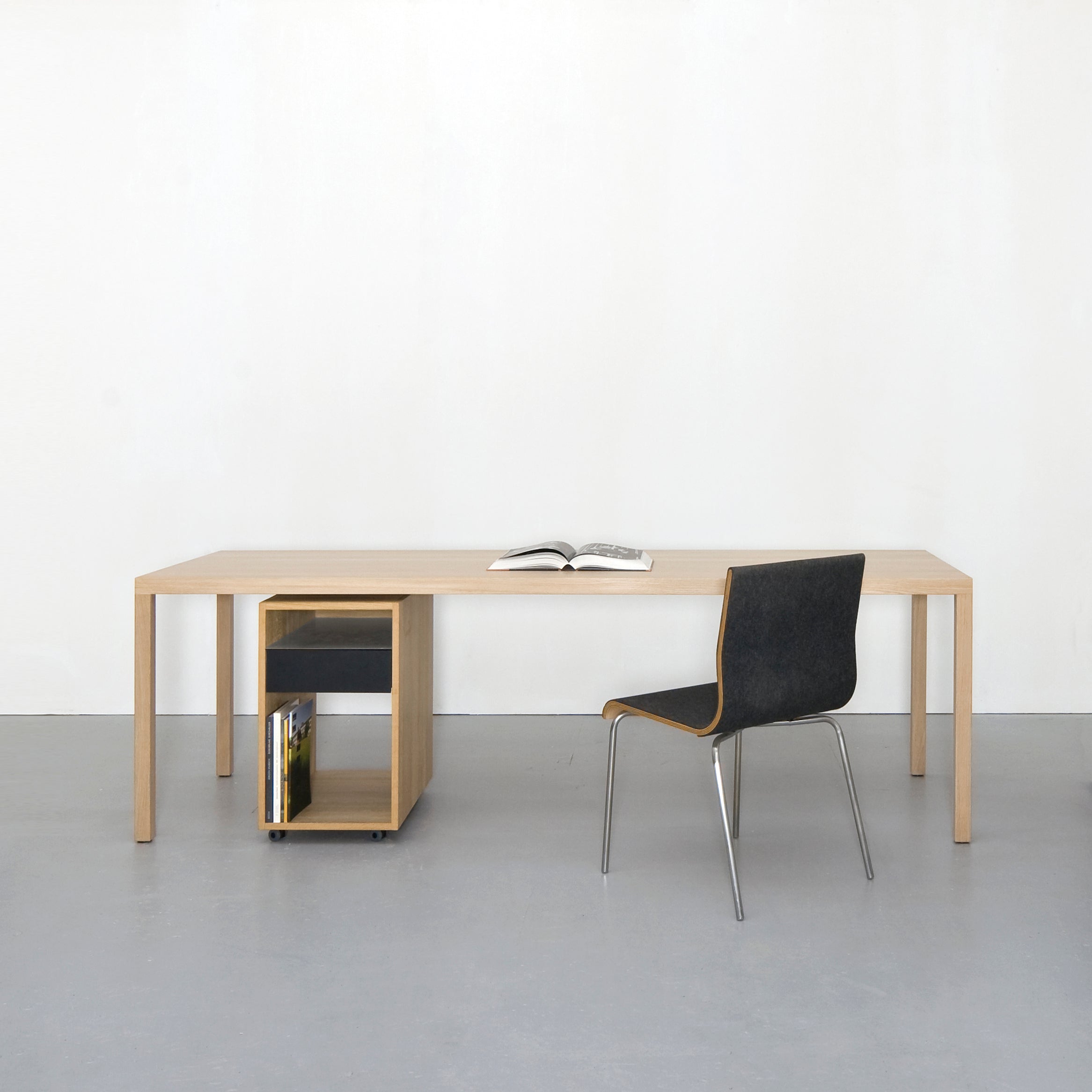 A minimalist workspace showcases a Sanktjohanser Dinavier Table with detachable legs alongside a modern chair on a gray floor. An open book rests on the table, while books are stored on a small shelf below. A plain white wall in the background completes the simple, clean aesthetic.