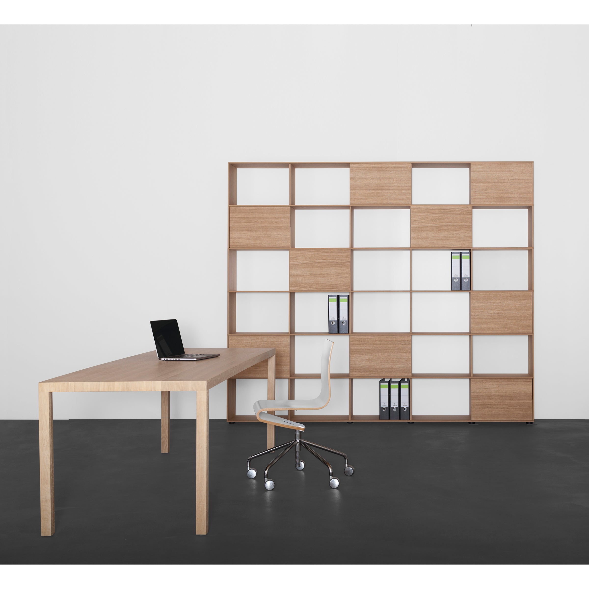 A minimalist office features a Sanktjohanser Dinanull Table as a wooden desk, an open laptop, and a white wheeled chair. A large wooden bookshelf with several shelves of folders stands against the white wall on a dark floor.