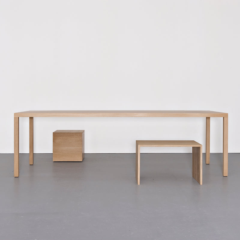 A minimalist scene showcases a Sanktjohanser Dinanull Table, a small wooden cube, and a wooden bench against a plain white wall. The smooth gray floor enhances the settings simplicity and elegance.