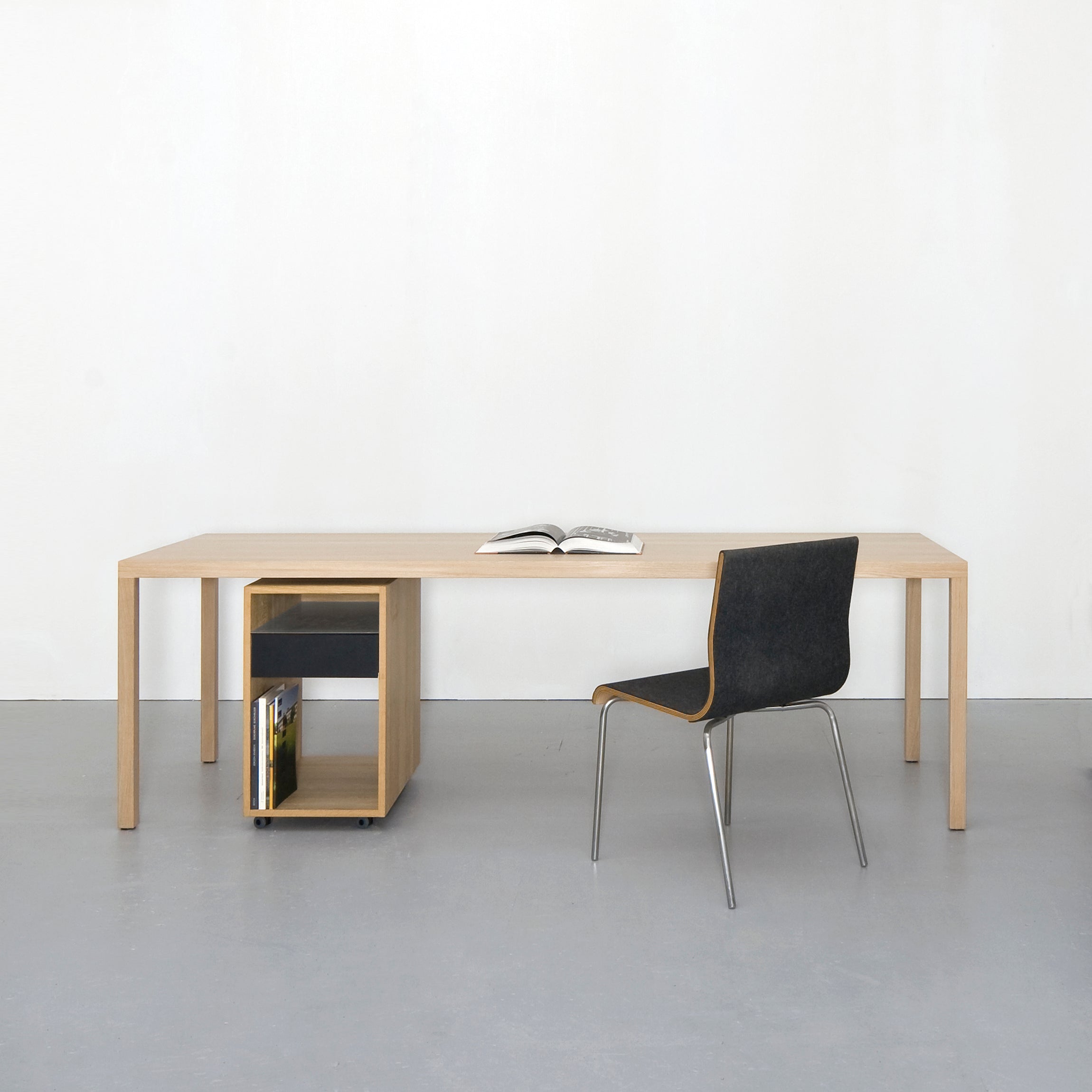 In a minimalist room, a Sanktjohanser Dinanull table pairs elegantly with a black chair featuring metal legs. An open book lies on the table, while below, a sleek shelf with black and wooden accents cradles books. The walls and floor maintain their neutral palette.