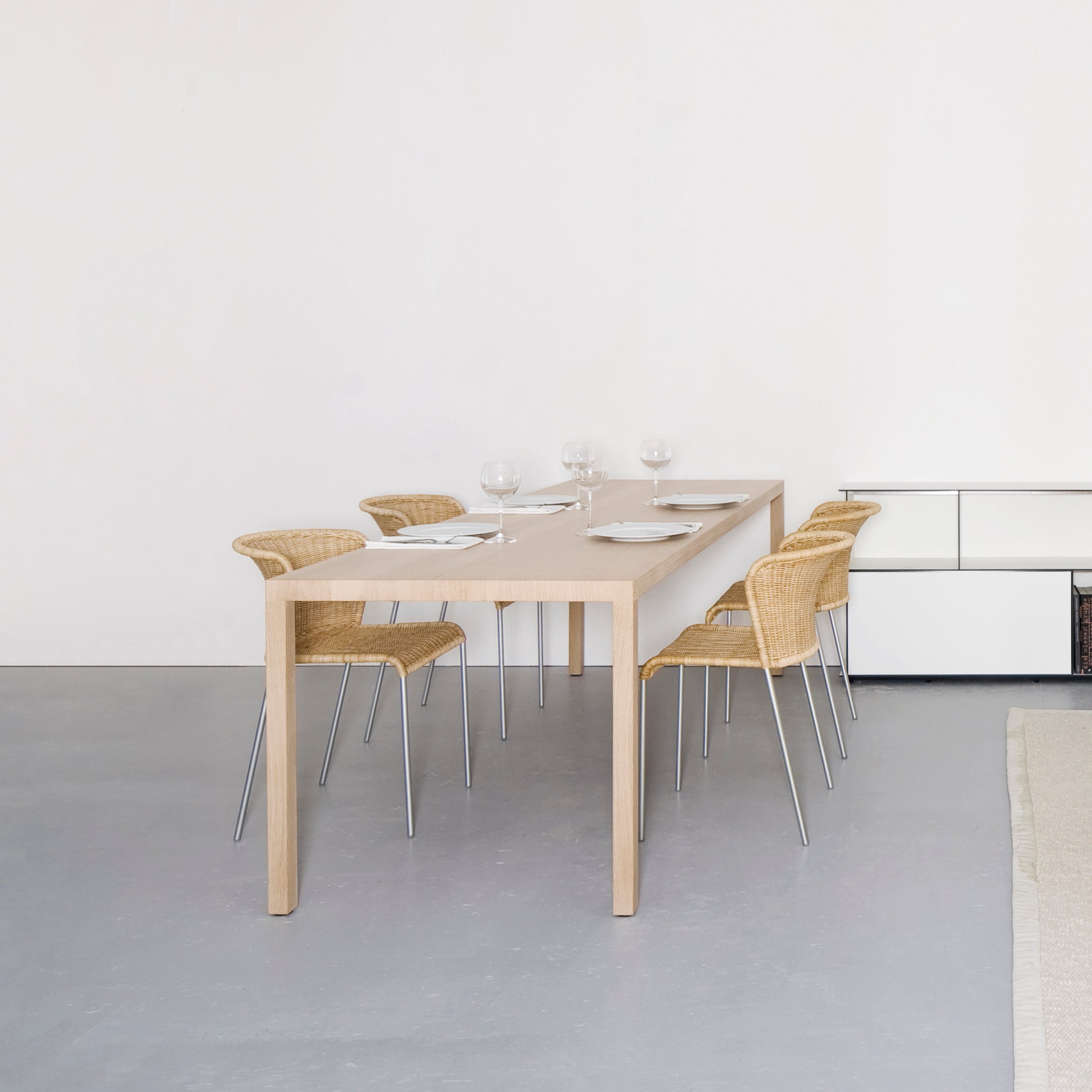 A minimalist dining room features the Dinanull Table by Sanktjohanser, a solid wood set for six perfectly paired with woven chairs. Adorned with white plates, glasses, and napkins, the bright and inviting space boasts a neutral palette and simple decor.