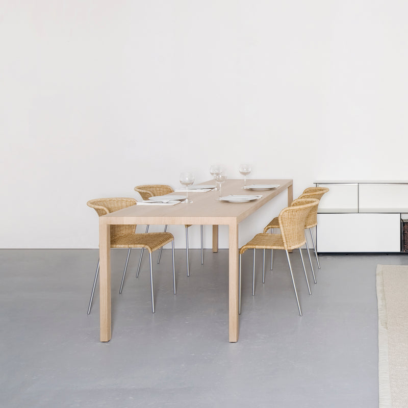 A minimalist dining room features the Dinanull Table by Sanktjohanser, a solid wood set for six perfectly paired with woven chairs. Adorned with white plates, glasses, and napkins, the bright and inviting space boasts a neutral palette and simple decor.