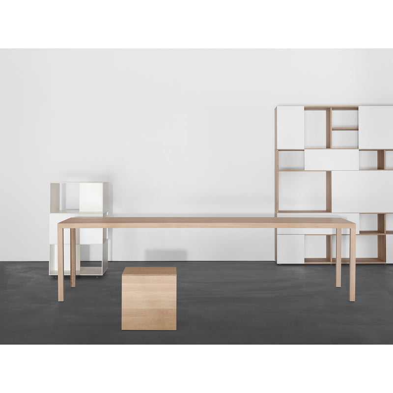 A minimalist room showcases a long, light-wood Sanktjohanser Dinanull Table and stool, complemented by modern geometric shelves with white and wood accents against a plain white wall. The dark grey floor adds depth to the serene space.