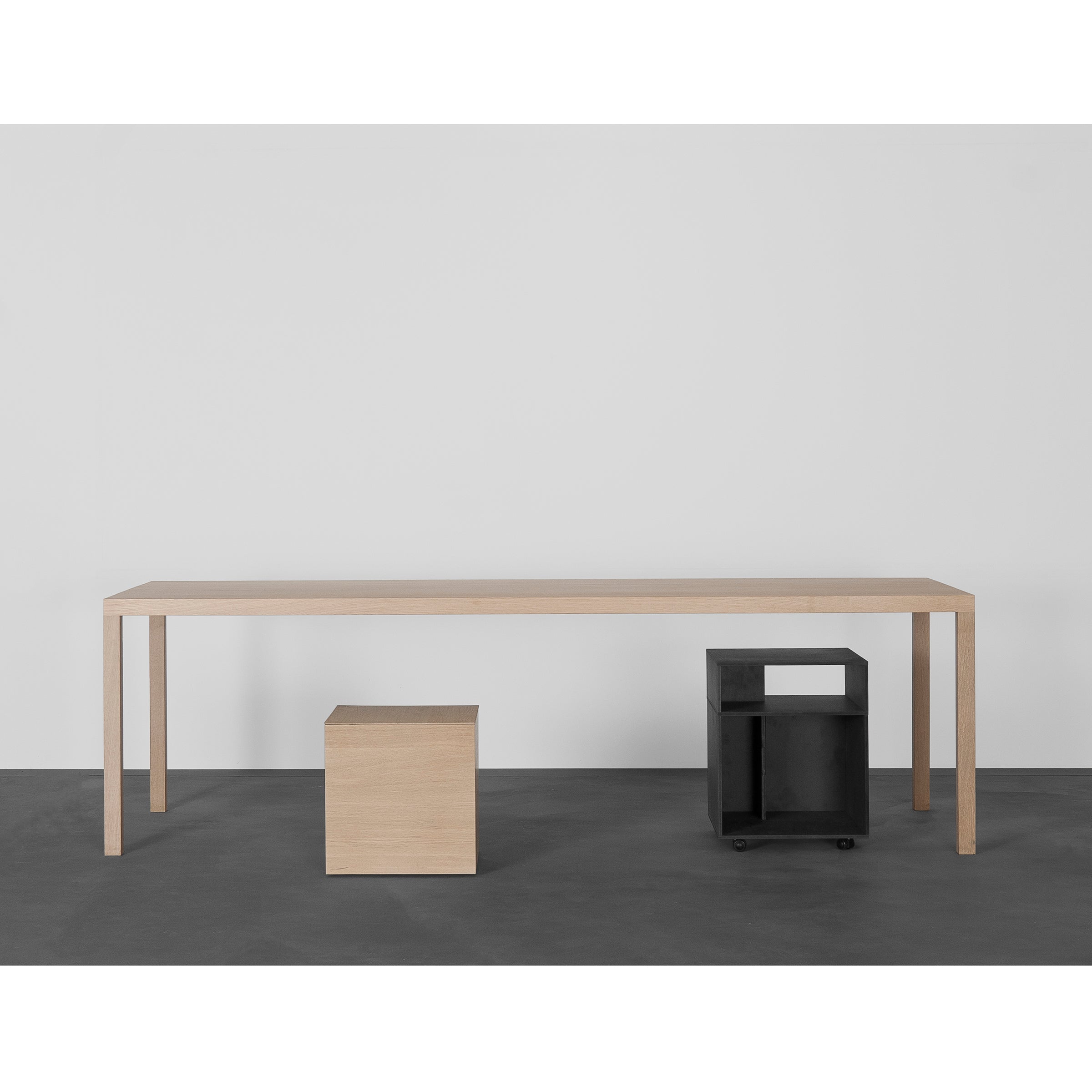 A minimalist room showcases the Sanktjohanser Dinanull Table with thin legs, a small wooden cube stool, and a black wheeled cart with an open compartment, all set against a plain white wall on a dark gray floor.