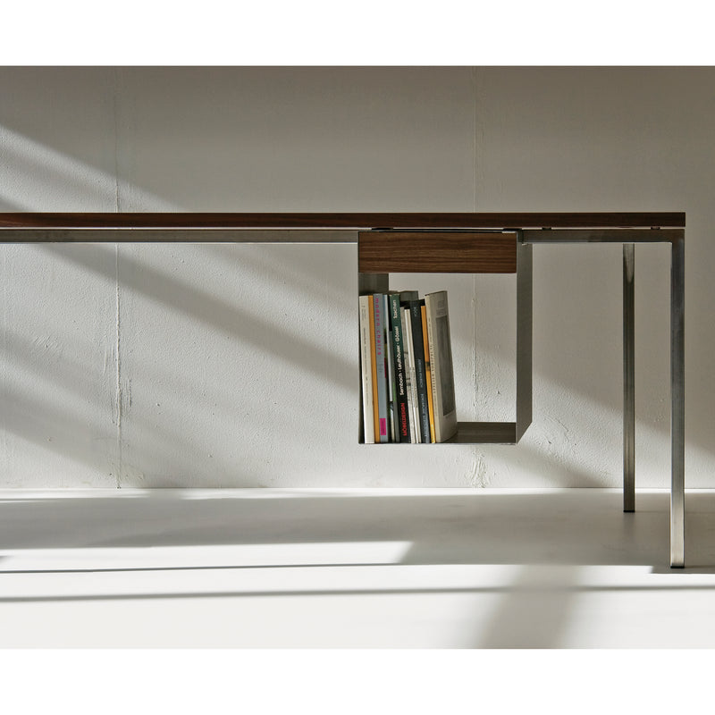 The Excenter Table by Sanktjohanser features a minimalist design with a solid wood top, thin metallic legs, and a unique integrated shelf for books. Soft shadows on white walls enhance its modern aesthetic, and stainless steel accents add elegance.
