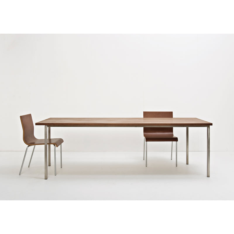 A minimalist dining area includes a Sanktjohanser Excenter Table made of solid wood with sleek stainless steel legs, accompanied by two brown chairs with metal frames. The simplicity is highlighted by a plain white wall and floor.