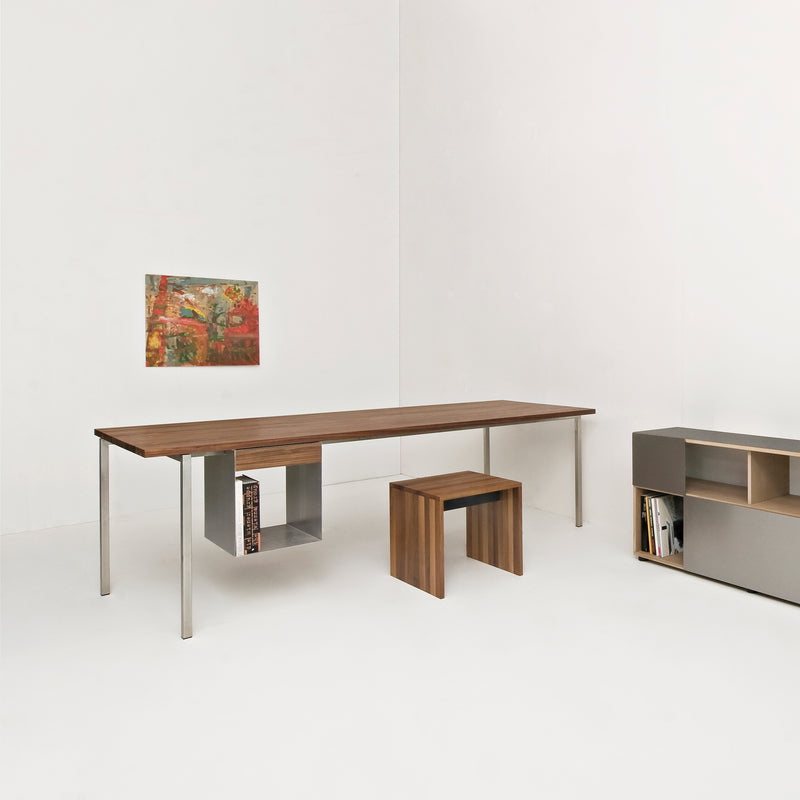 A minimalist room showcases the Excenter Table by Sanktjohanser, featuring solid wood with sleek stainless steel legs. Its paired with a matching stool and enhanced by a small abstract painting on the white walls. On the right, a modern low shelf holds books, adding to the serene ambiance.