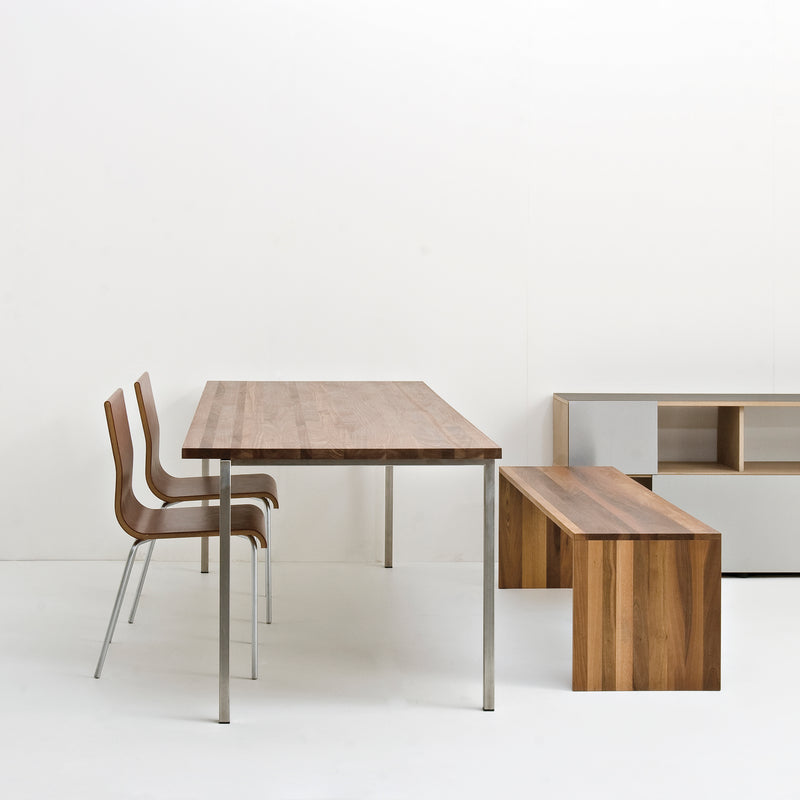 Sanktjohansers Excenter Table, crafted from solid wood with stainless steel legs, is part of a minimalist dining set with two sleek wooden chairs and a matching bench. A white modern shelving unit enhances the setting against a plain white wall.