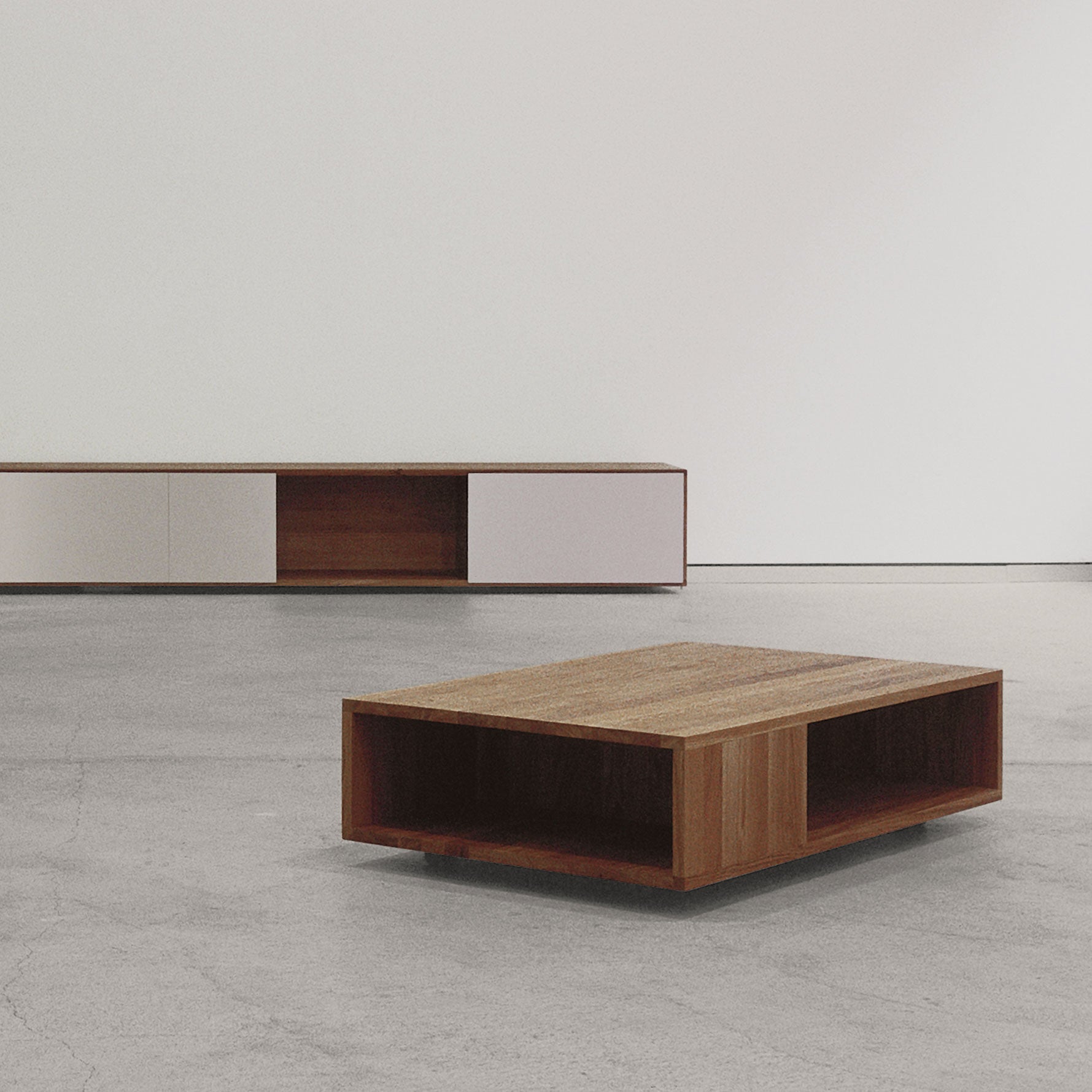 A minimalist room features two solid wood pieces on a smooth, gray concrete floor. A Sanktjohanser Flat coffee table with open sides sits in the foreground. Against the wall is a low sideboard with white doors, creating a modern and simple look.