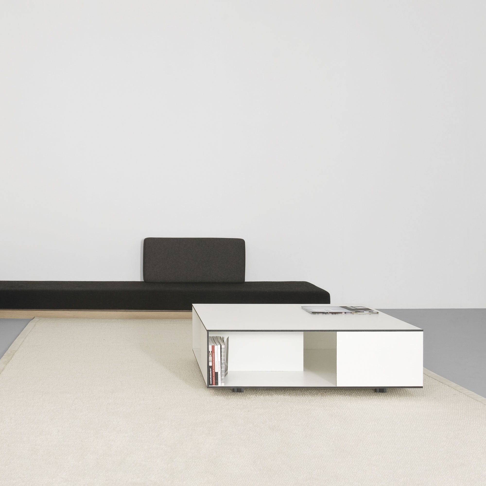 A minimalist room boasts a dark green bench against a white wall, enhanced by solid wood compartment shelves. A Sanktjohanser Flat coffee table sits on a light beige rug, adorned with books and magazines, maintaining the rooms simple, clean design.