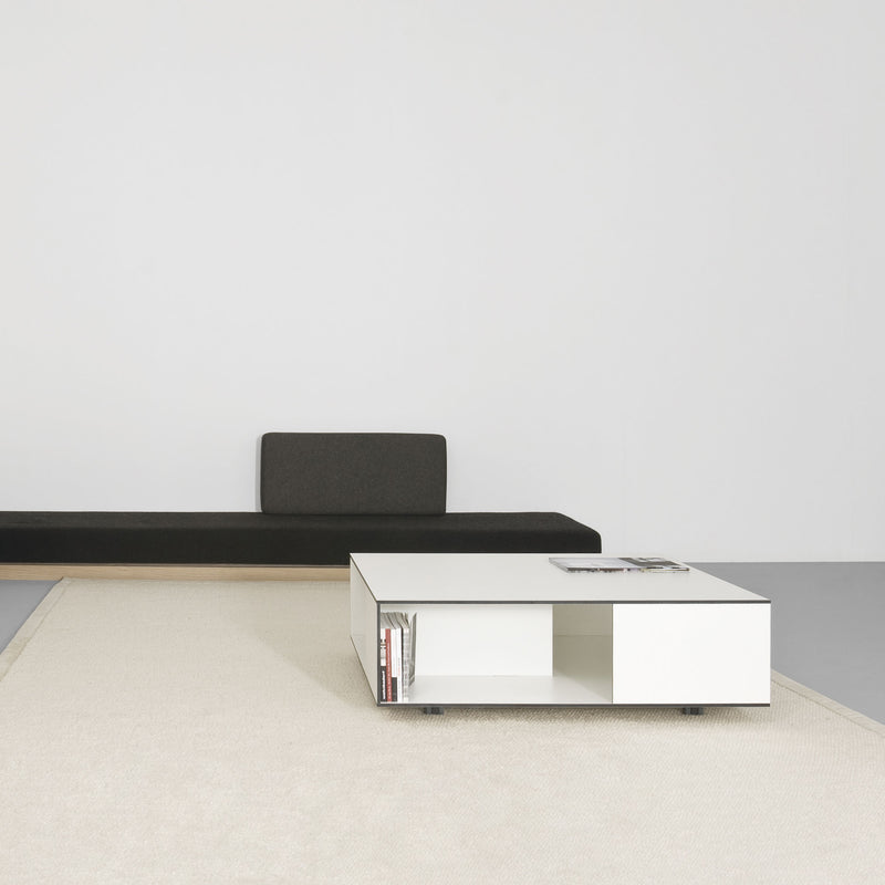 A minimalist room boasts a dark green bench against a white wall, enhanced by solid wood compartment shelves. A Sanktjohanser Flat coffee table sits on a light beige rug, adorned with books and magazines, maintaining the rooms simple, clean design.