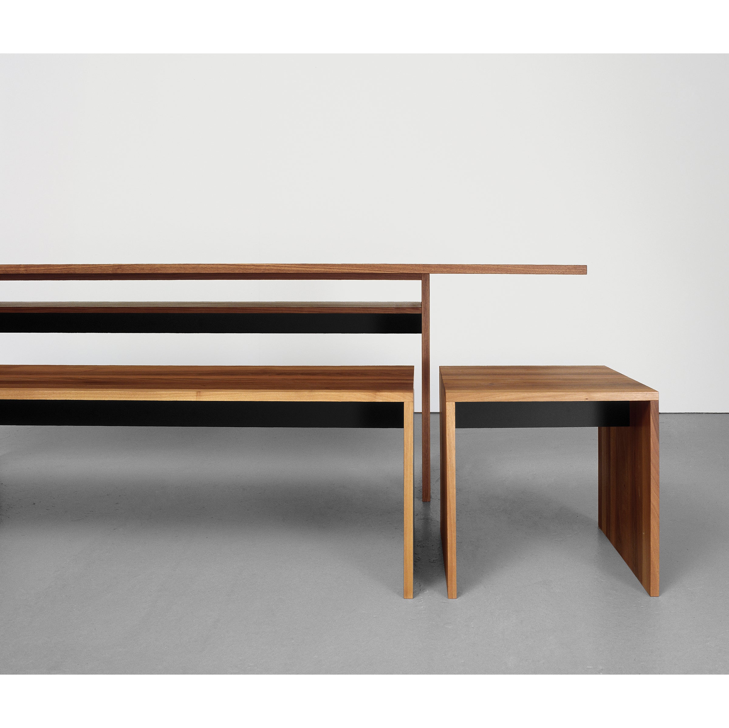 The Sanktjohanser Format Bench, part of a minimalist dining set made from solid wood, includes a long rectangular table and a smaller square table with sleek lines. The set is showcased on a smooth light gray floor against a plain white wall.
